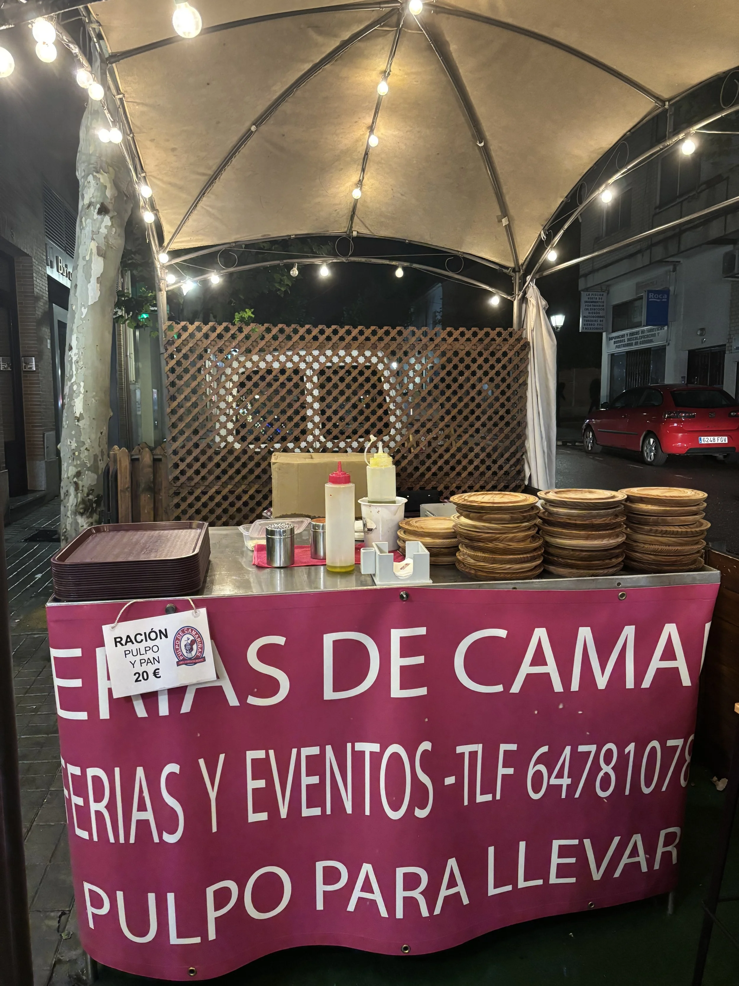 Carrito de comida con platos de madera apilados, botellas de salsa y un cartel que dice: "Ración pulpo y pan 20€". Está en un puesto en la calle con luces y una lona.