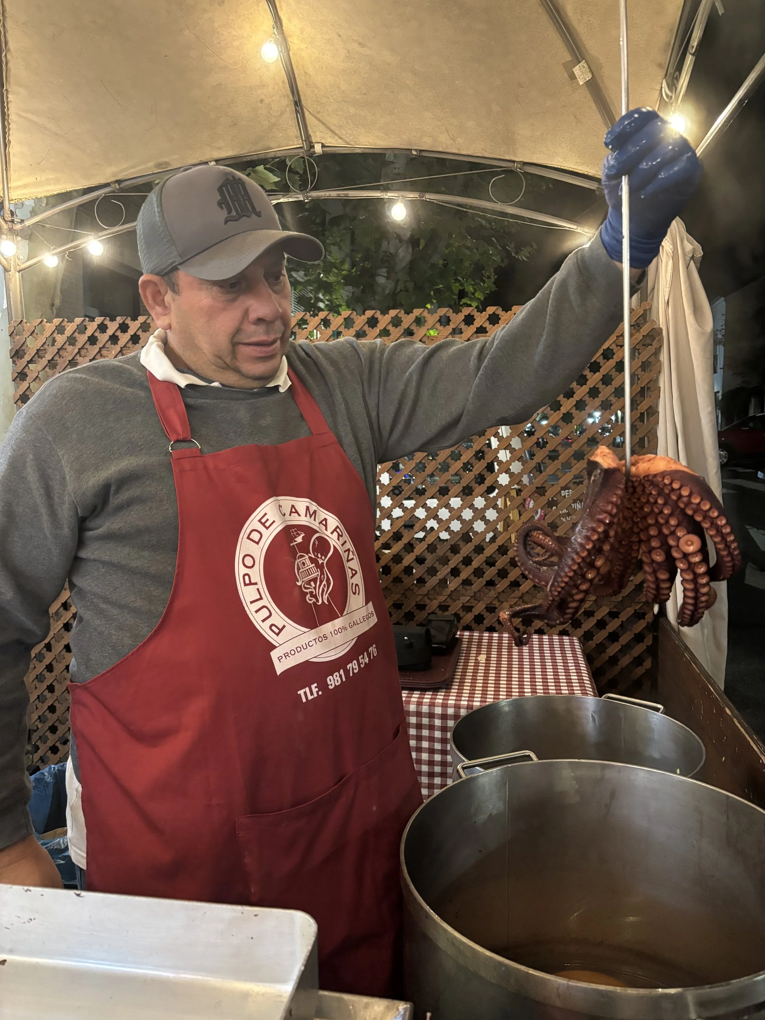 Un hombre cocinero sosteniendo un pulpo cocido en un pincho en un puesto de comida, usando un delantal rojo y un gorro gris, en un evento al aire libre.