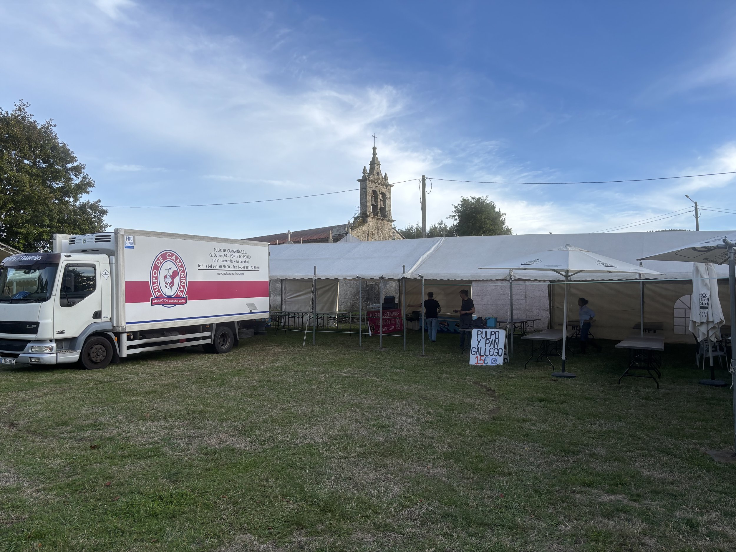 Carpa de comida en un campo, con un camión de transporte y un cartel que anuncia pesca y pan gallego por 15 euros. En el fondo, una iglesia y árboles, cielo despejado.