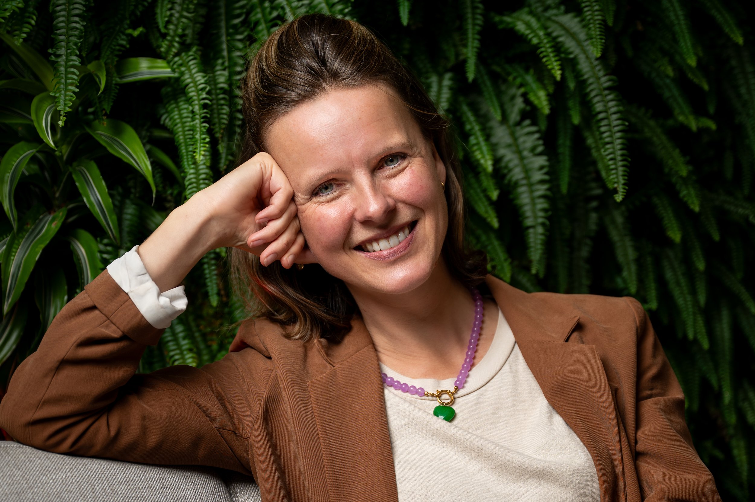 Een lachende vrouw met bruin haar, gekleed in een bruine jas en een witte top, zit in een groene omgeving met veel planten.