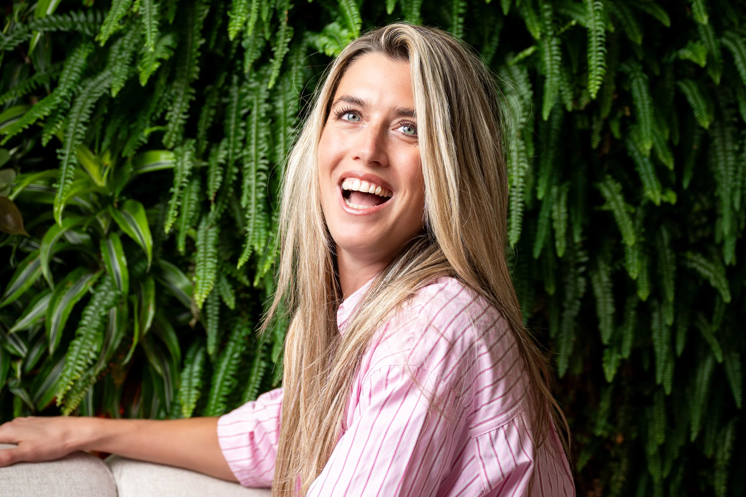Vrouw met lange blonde haren glimlacht in een tuin met groene varens.