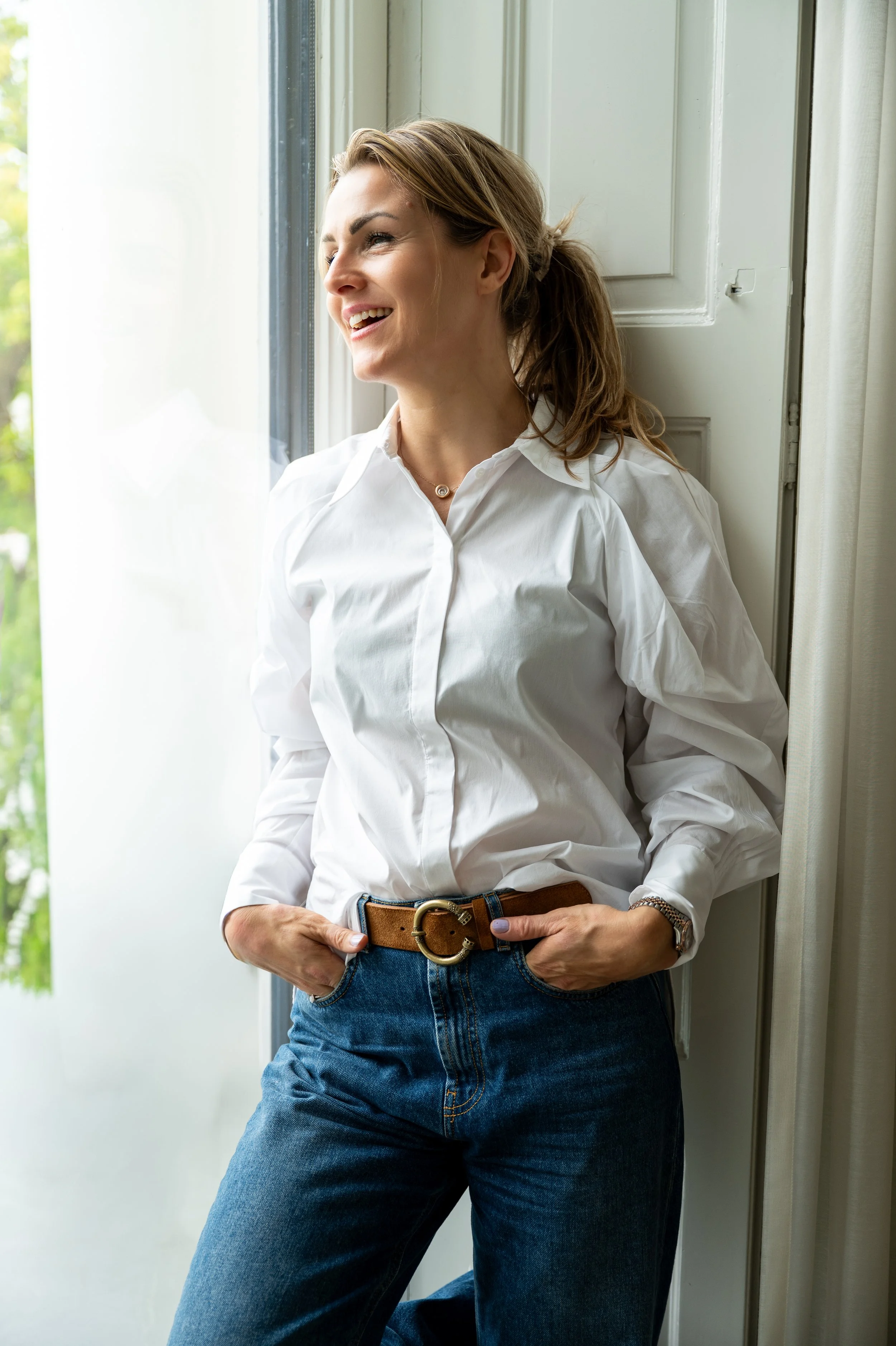 Een vrouw met een witte blouse en blauwe jeans leunt tegen een raam en glimlacht.