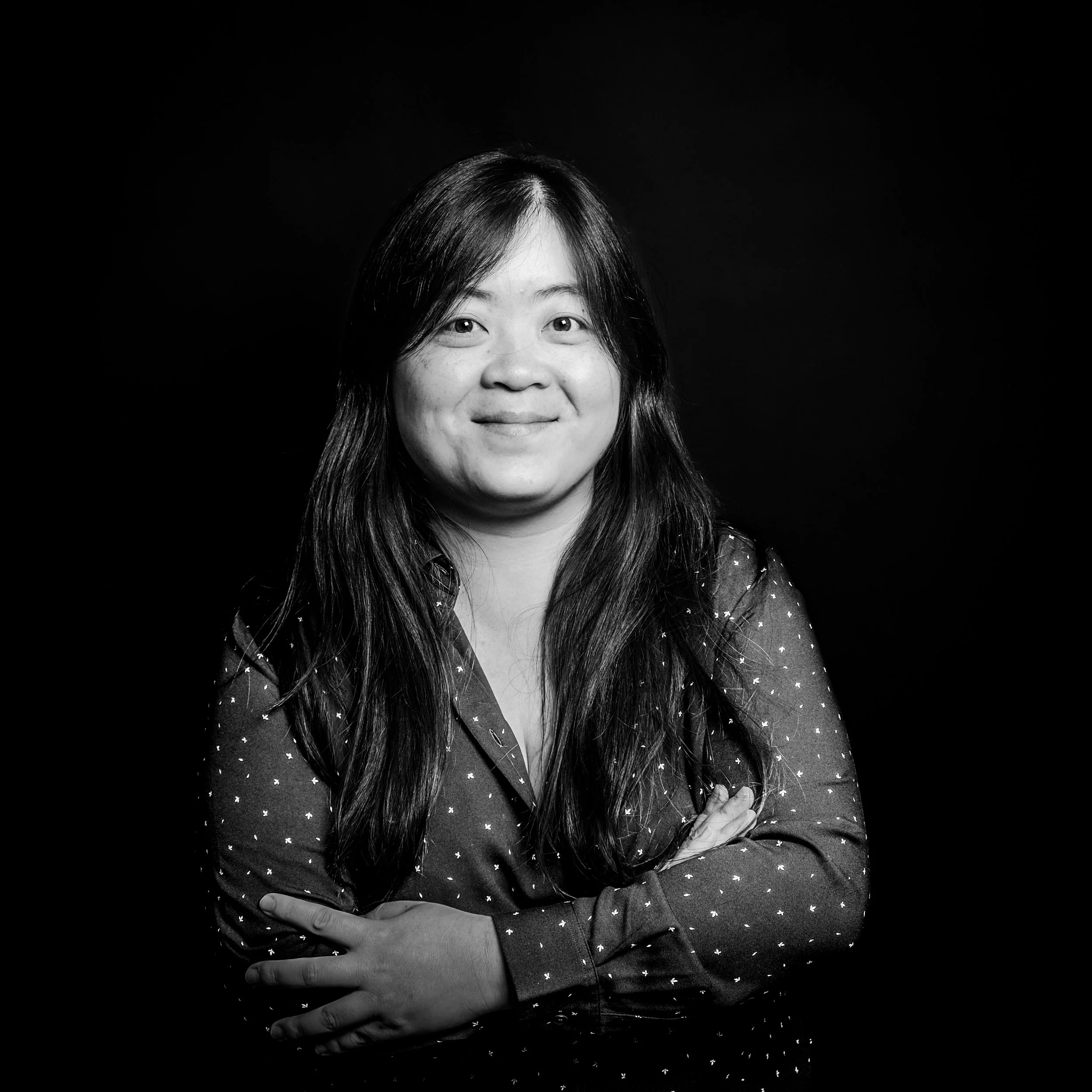 Black and white portrait of a woman with long hair, wearing a polka dot shirt, smiling with crossed arms.