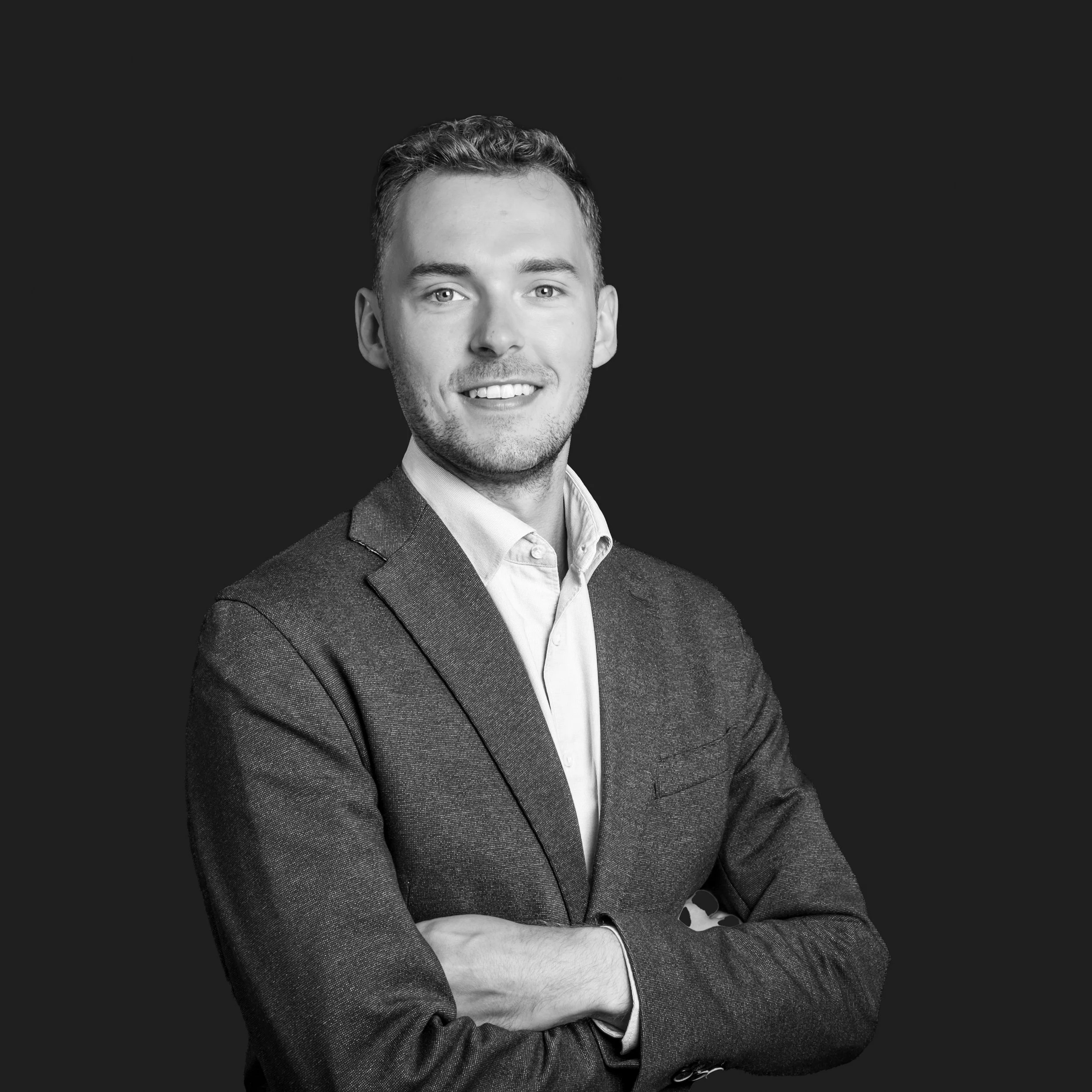 Black and white portrait of a young man with short hair, smiling, wearing a suit jacket and shirt, arms crossed, against a dark background.