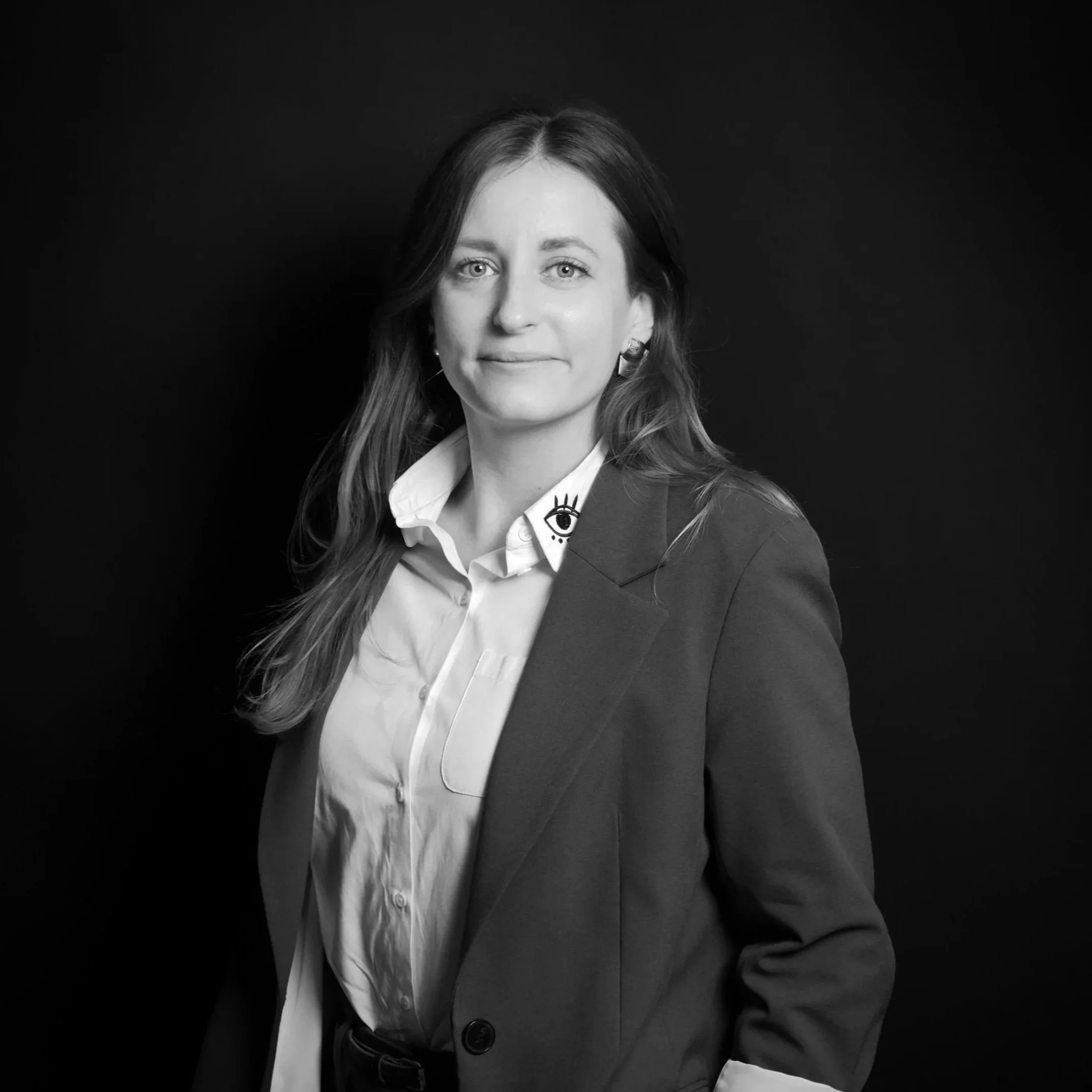 Black and white portrait of a woman with long hair wearing a blazer with a graphic eye design on the collar, and a white shirt, against a dark background.