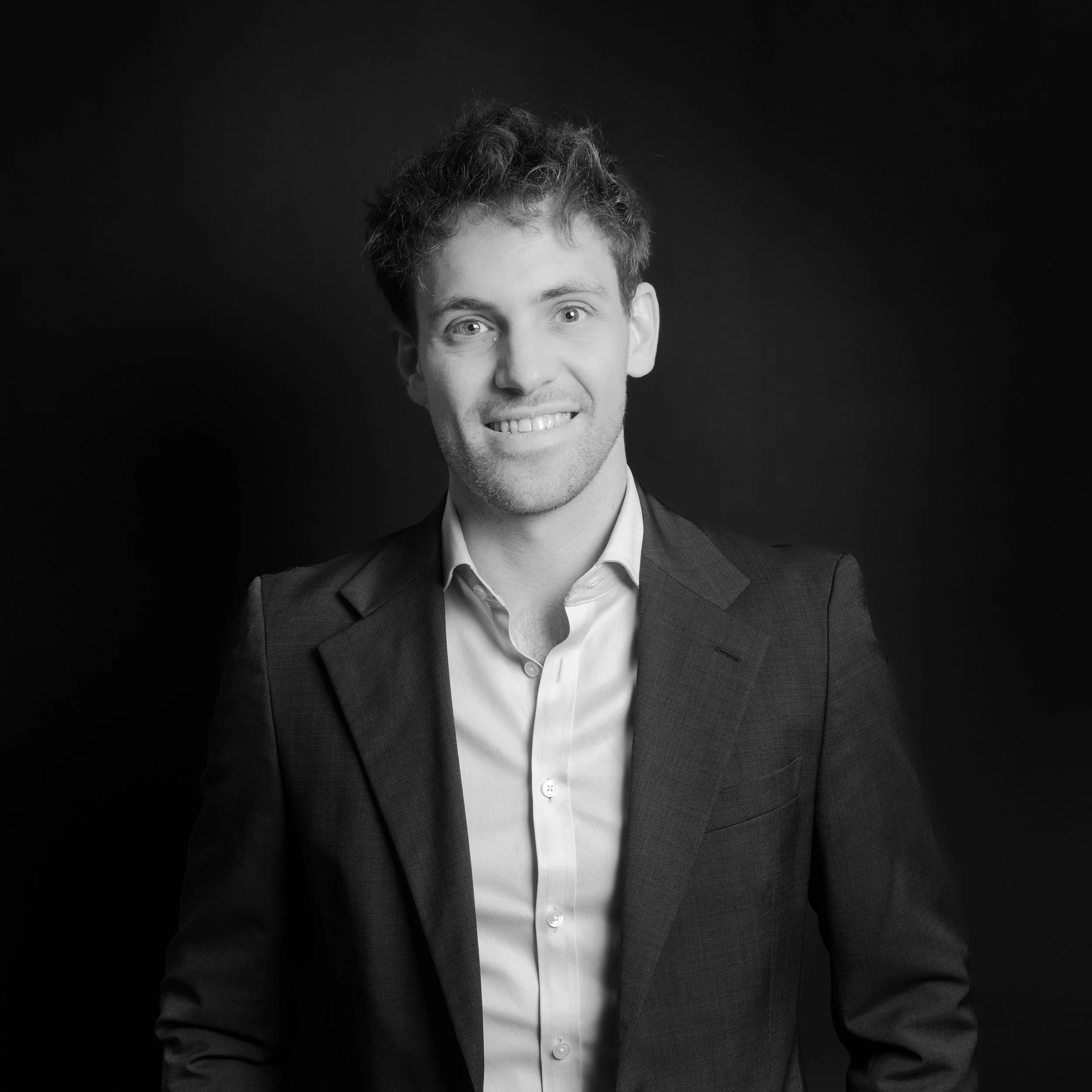 Black and white portrait of a smiling young man in a suit against a dark background.