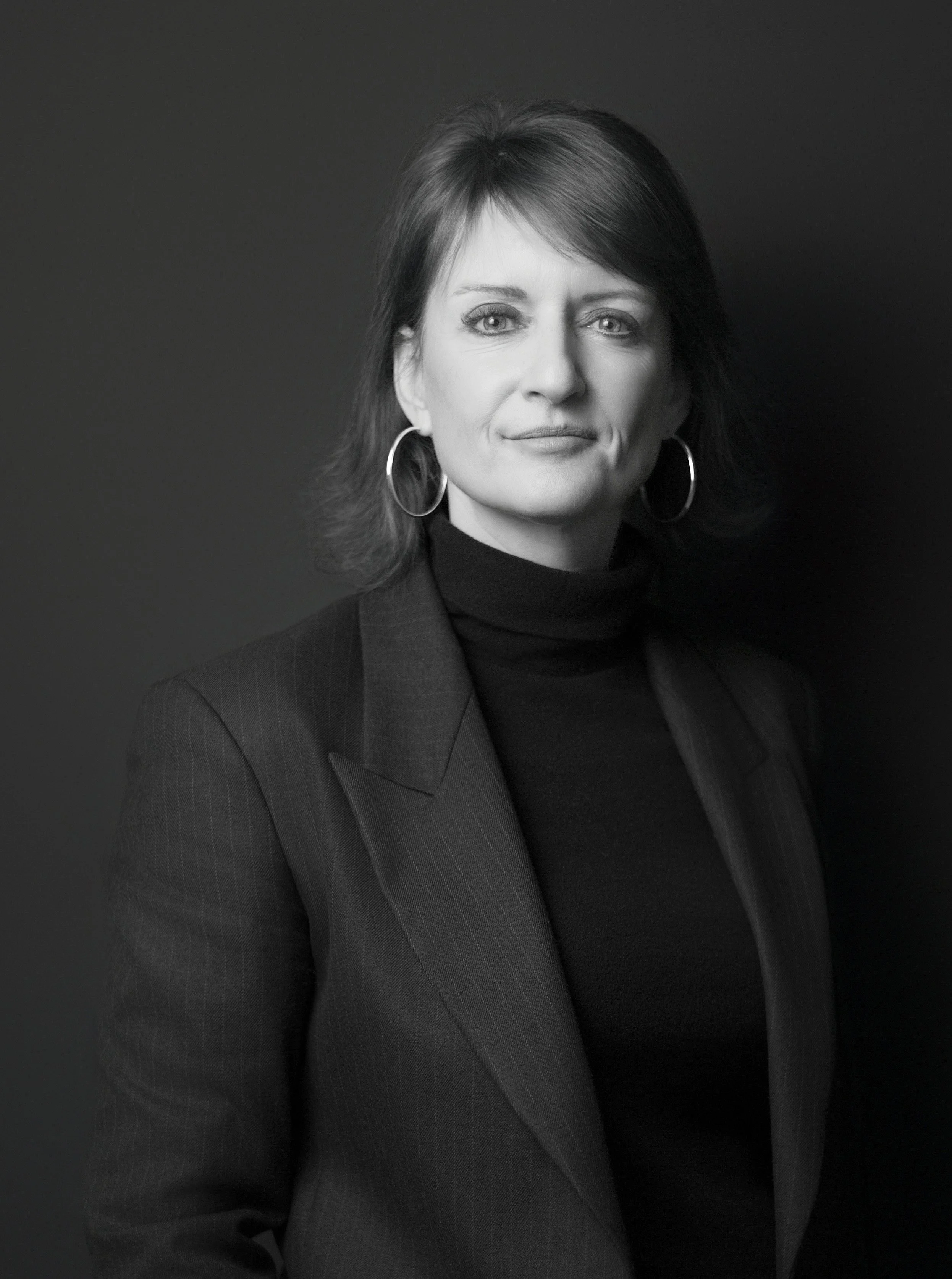 Black and white portrait of a woman with shoulder-length hair, wearing a blazer and a t-shirt with a logo, standing with arms crossed against a dark background.