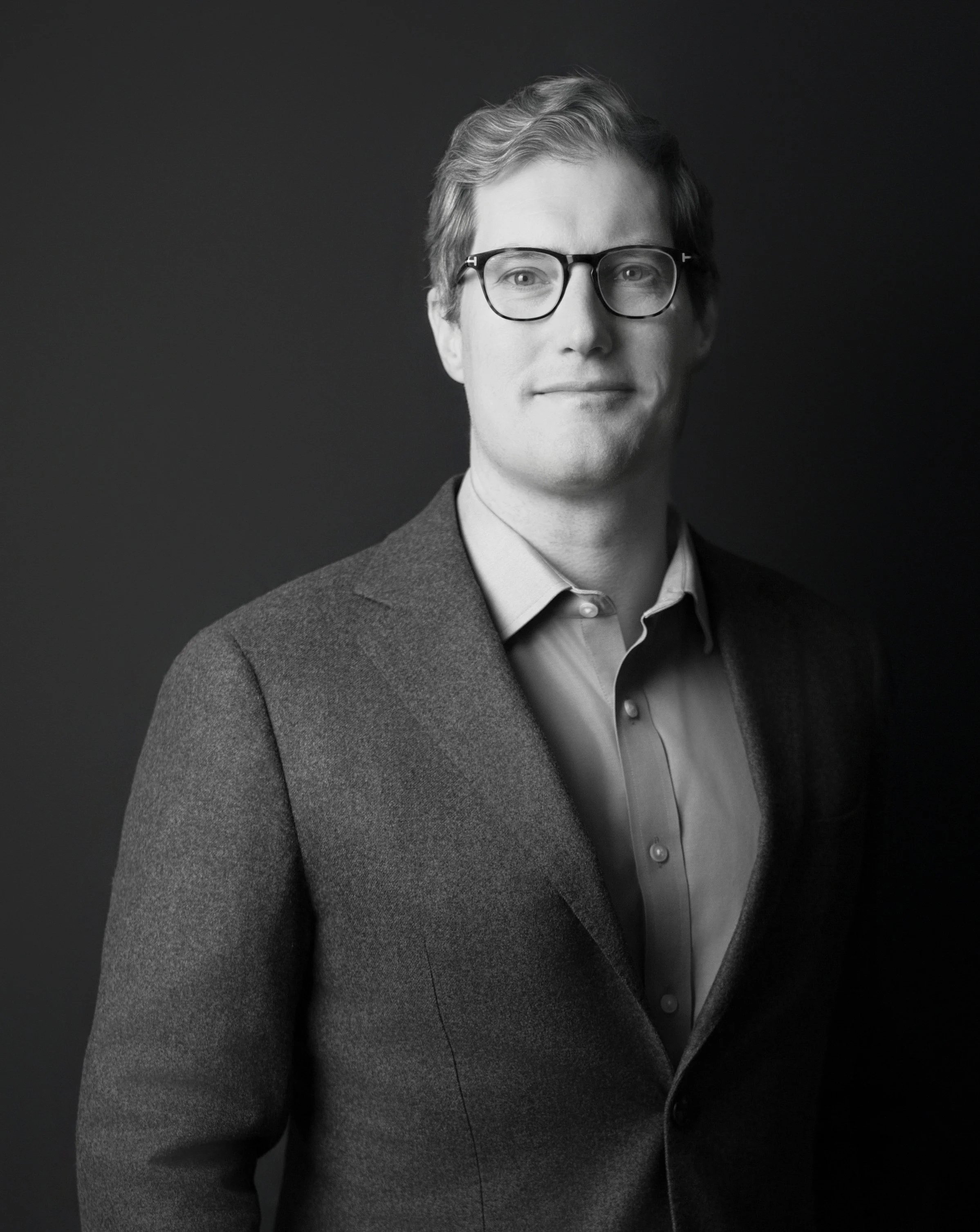Black and white portrait of a man with light hair, wearing a dark blazer over a white shirt, standing against a dark background with arms crossed.