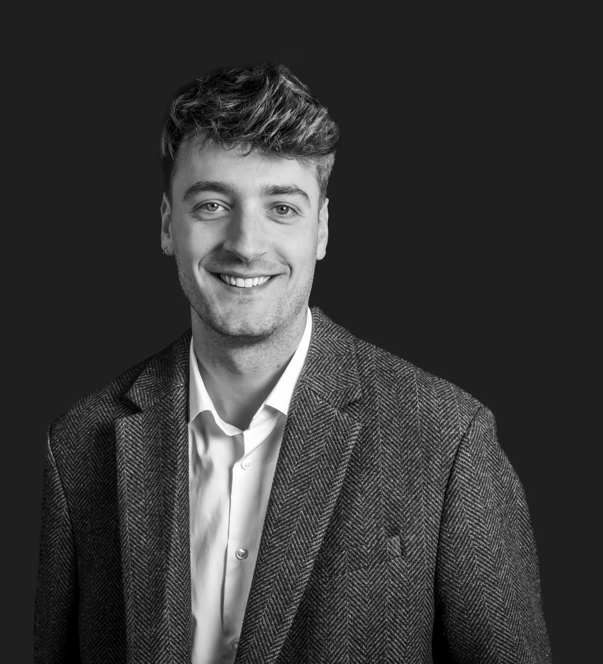 Black and white portrait of a young man with light hair, smiling, wearing a white shirt and a herringbone blazer against a dark background.