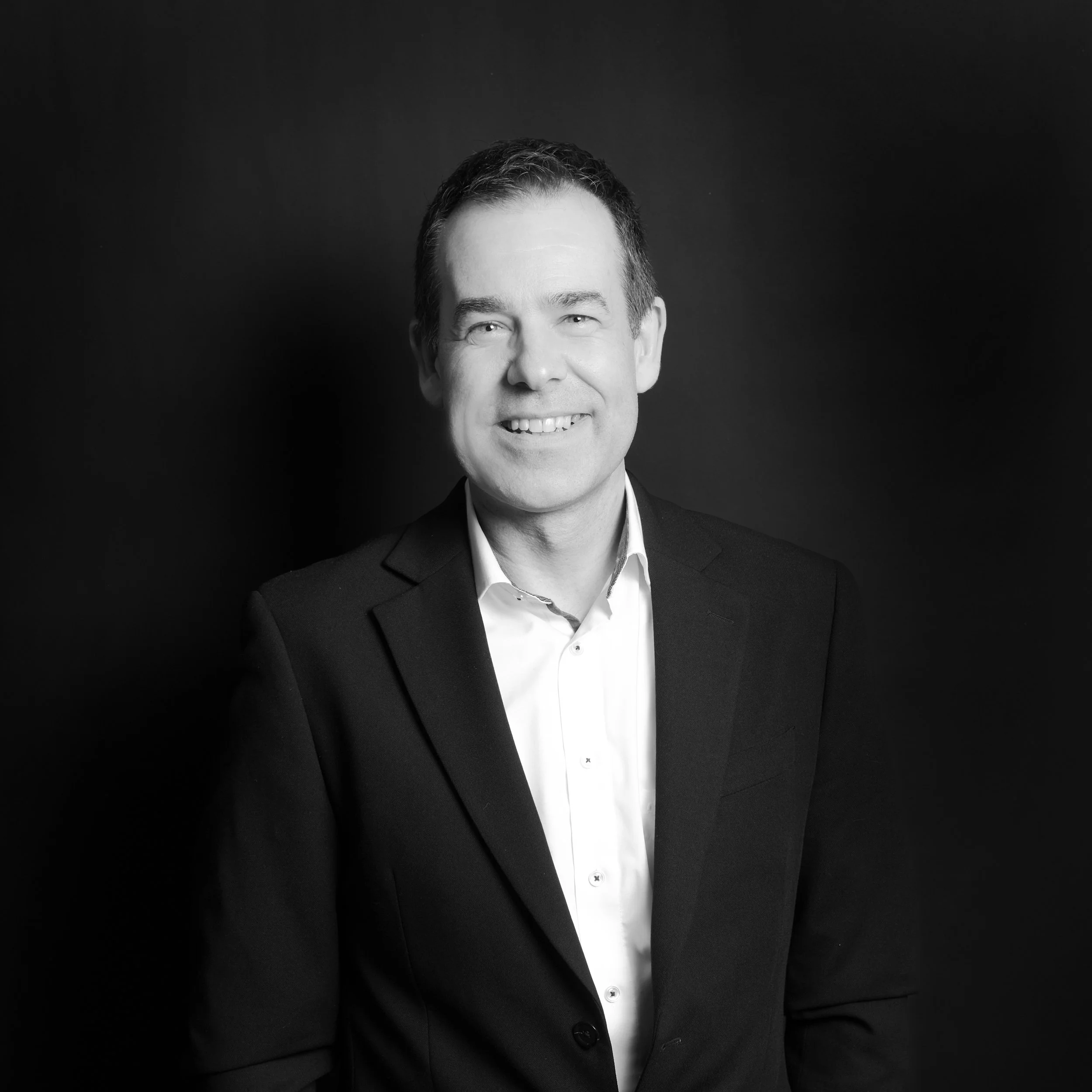 Black and white portrait of a smiling man in a suit and white shirt against a dark background.