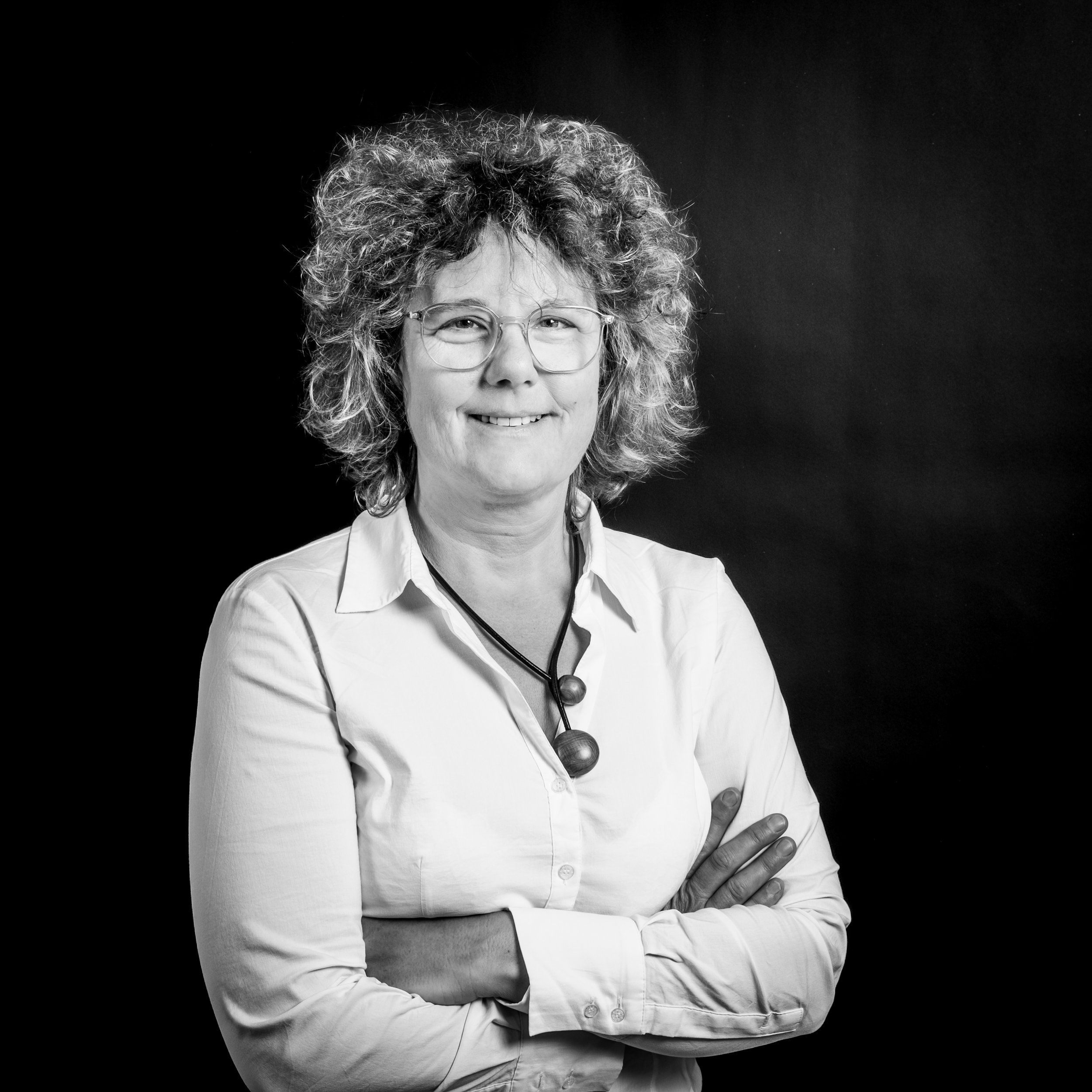 Black and white photo of a woman with curly hair and glasses, smiling, wearing a white shirt, with arms crossed, against a dark background.