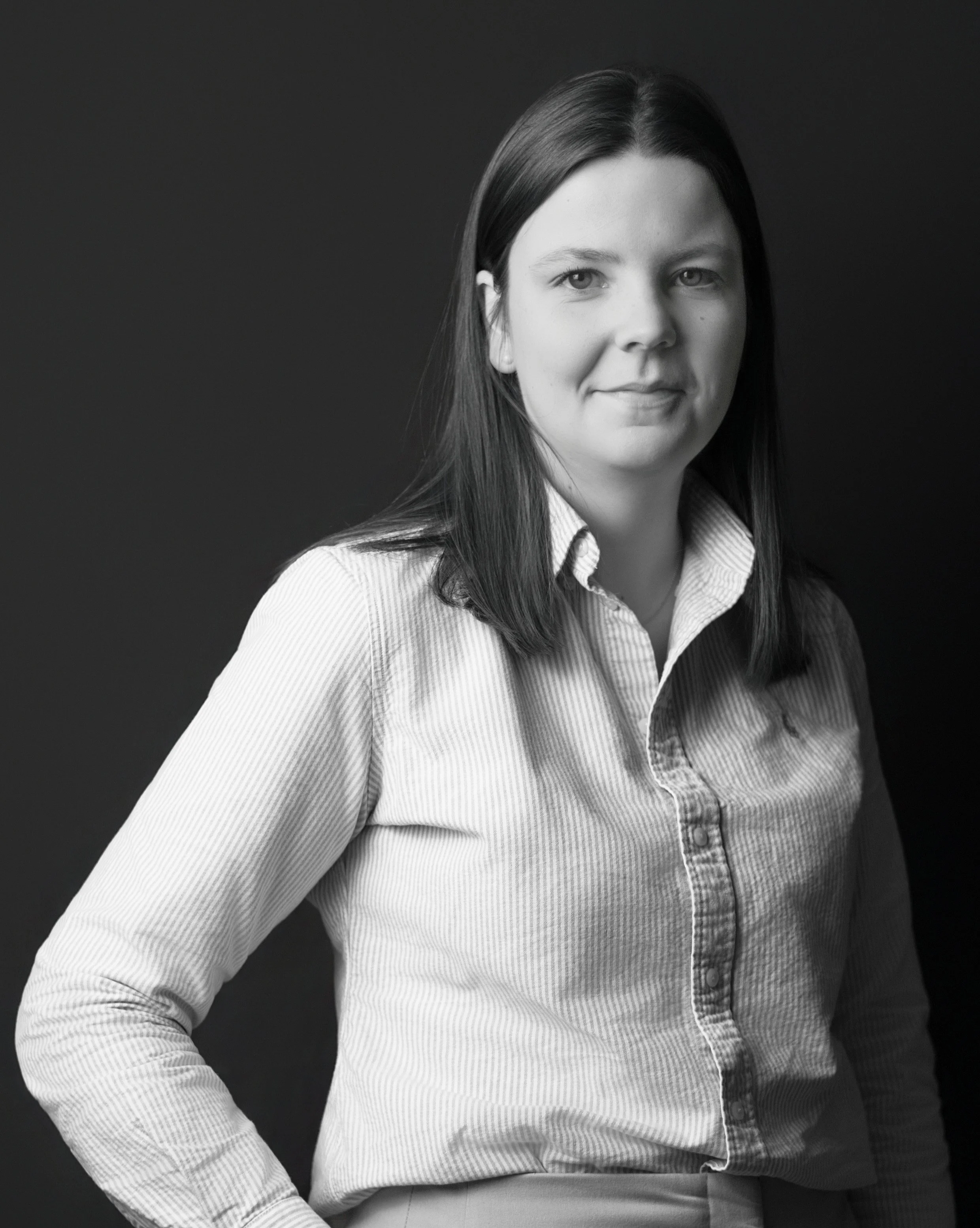 Black and white portrait of a woman with shoulder-length dark hair, wearing a short-sleeved button-up shirt, an Apple Watch, and crossed arms, against a dark background.