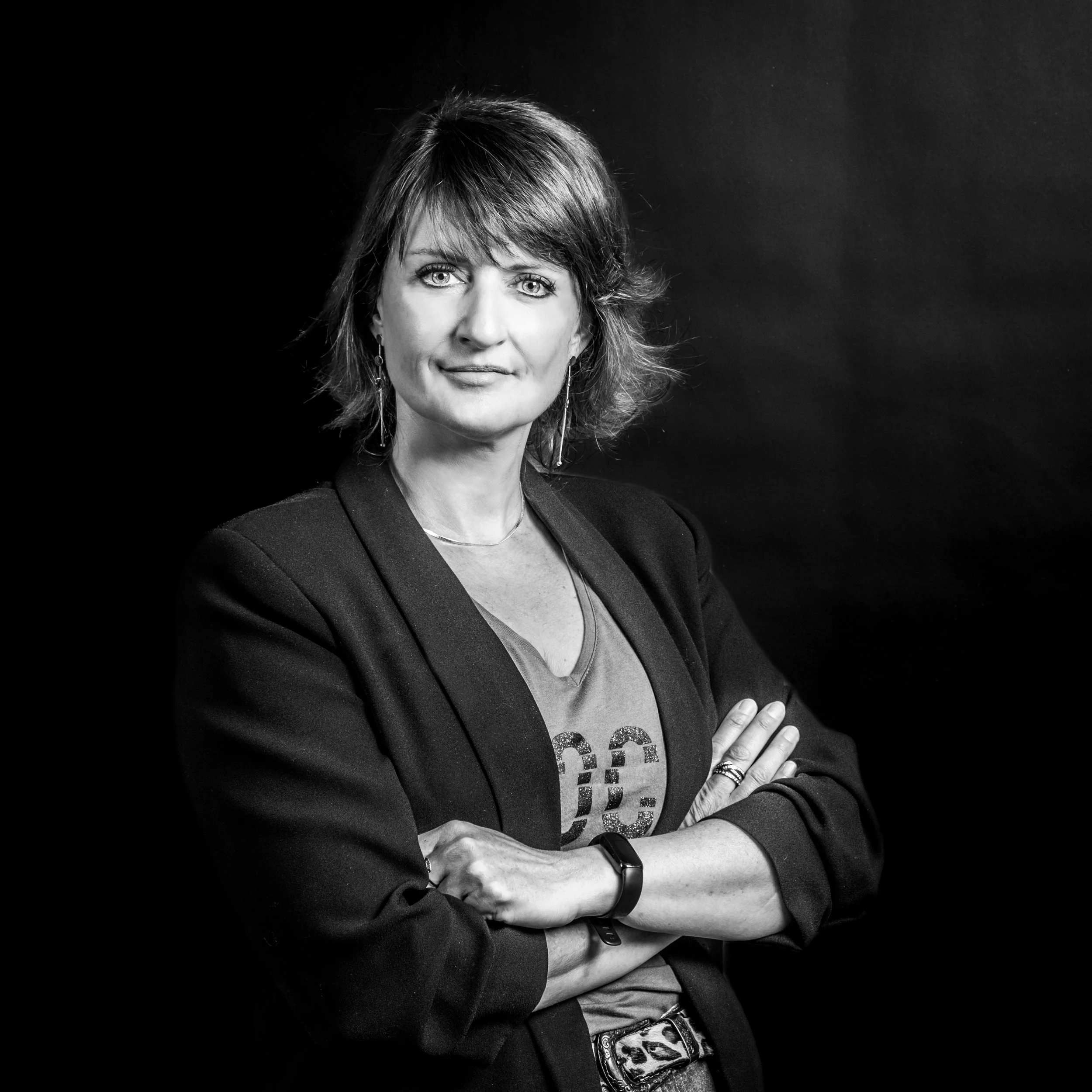Black and white portrait of a woman with shoulder-length hair, wearing a blazer and a t-shirt with a logo, standing with arms crossed against a dark background.