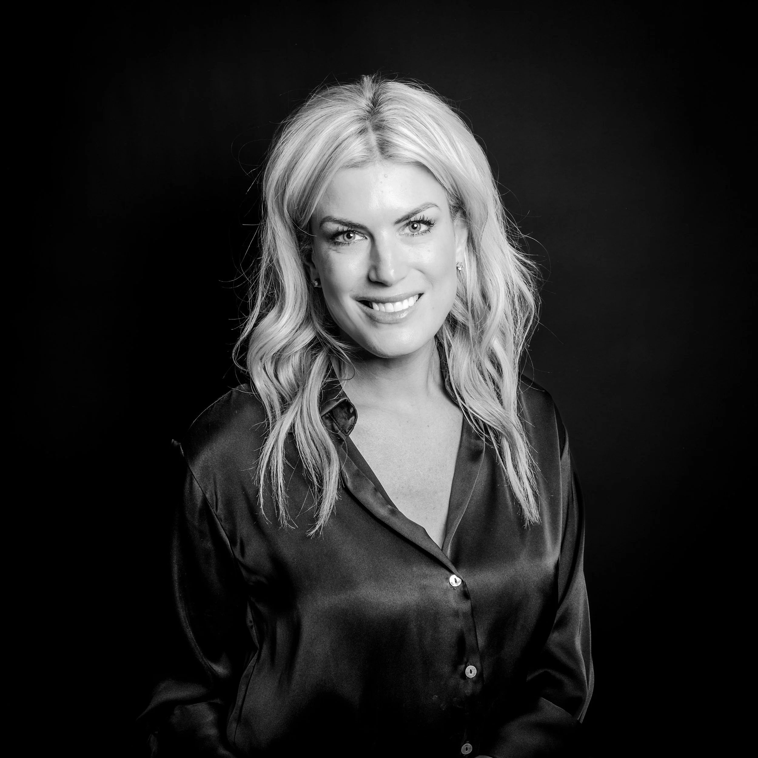 Black and white portrait of a smiling woman with wavy blonde hair, wearing a satin blouse, against a dark background.