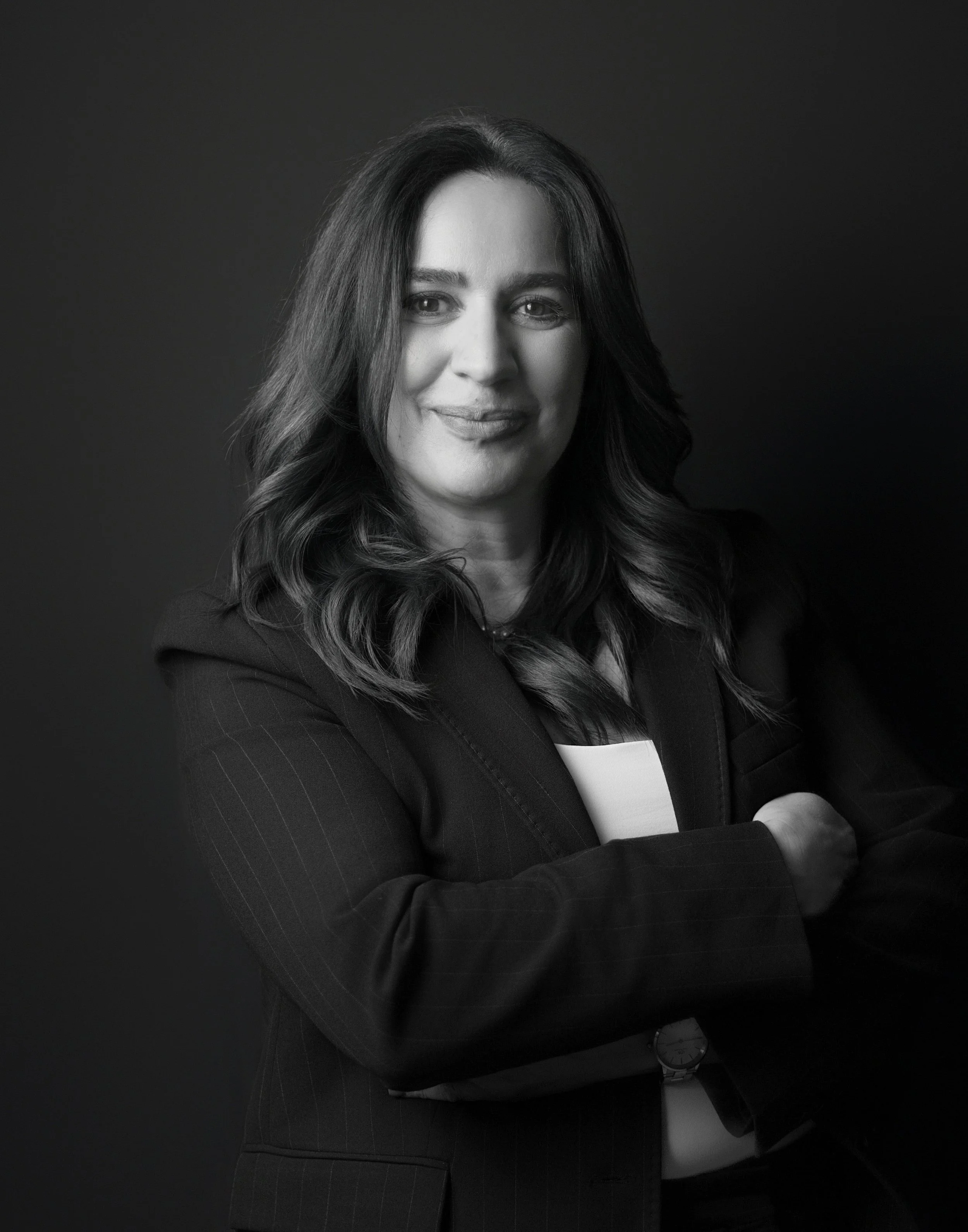Black and white portrait of a woman with shoulder-length wavy hair, wearing glasses, a blazer, and a blouse, smiling against a plain black background.