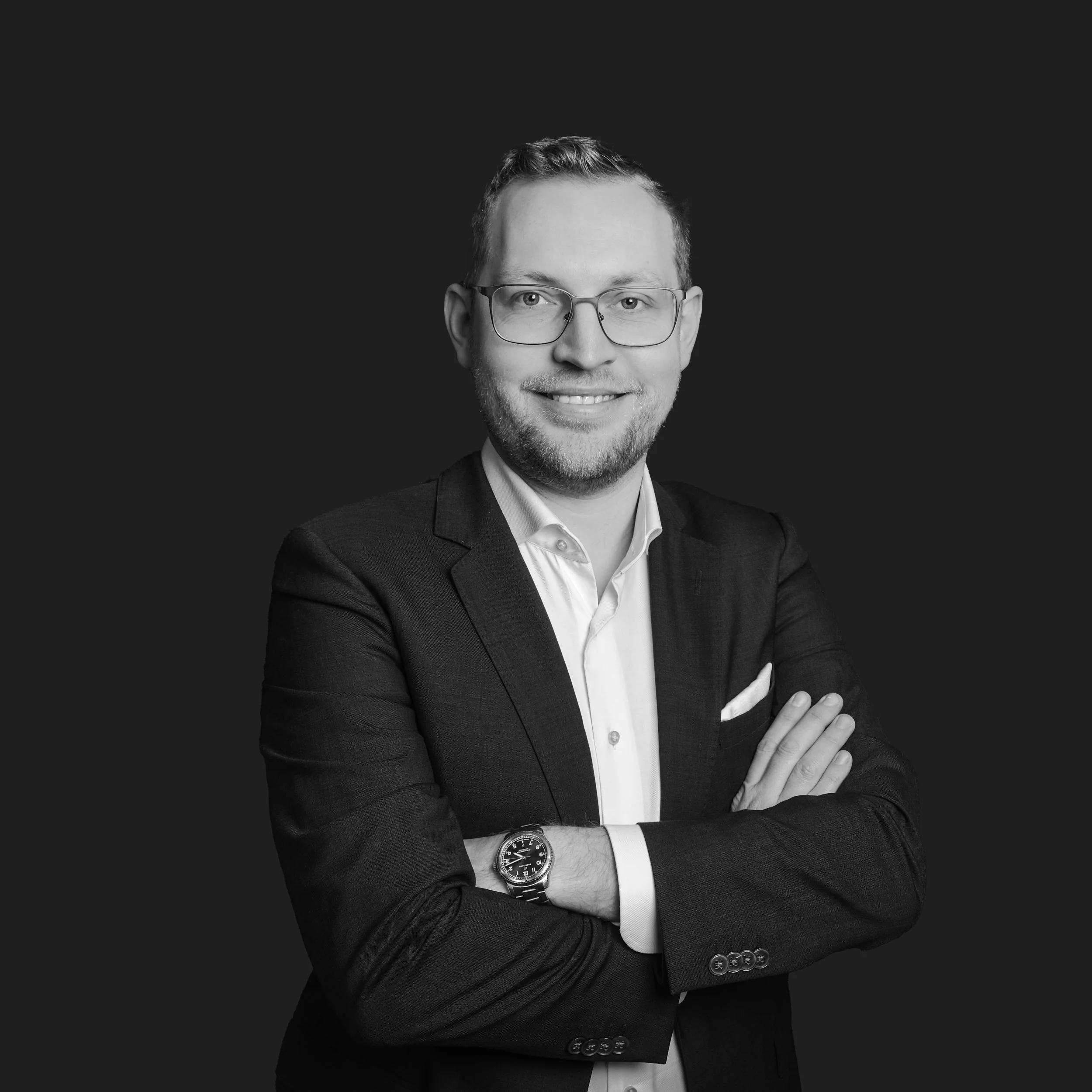 Black and white portrait of a man wearing glasses, a suit jacket, white shirt, crossed arms, and a wristwatch, smiling against a dark background.