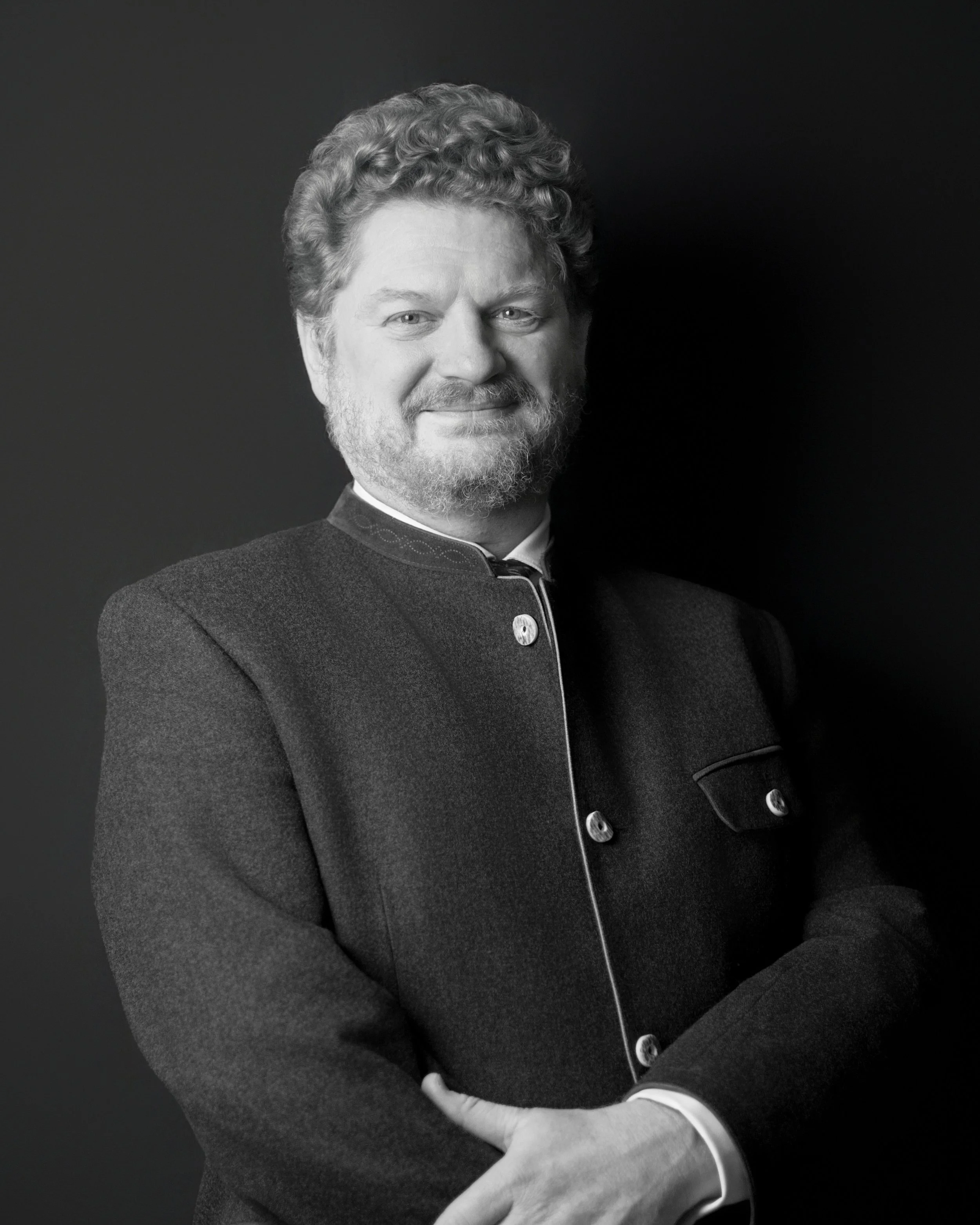Black and white portrait of a smiling man with glasses, curly hair, and a beard, wearing a dark velvet blazer and a light-colored shirt, standing with arms crossed against a black background.