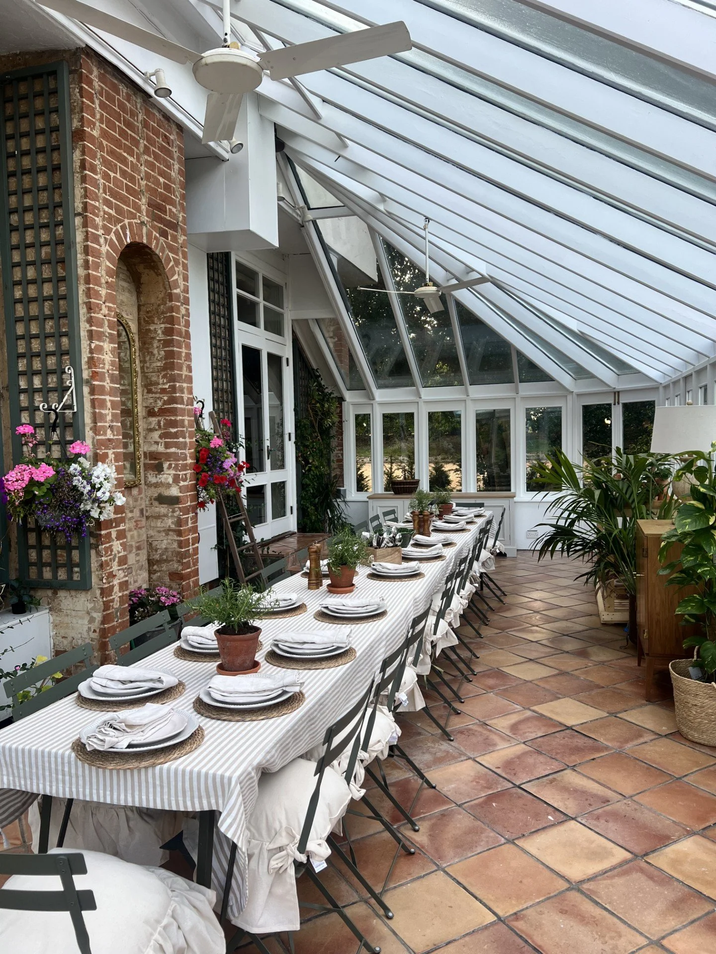 A long dining table set for a meal in a sunroom with glass walls and ceiling, decorated with potted plants, plates, and utensils, with chairs neatly arranged around it.