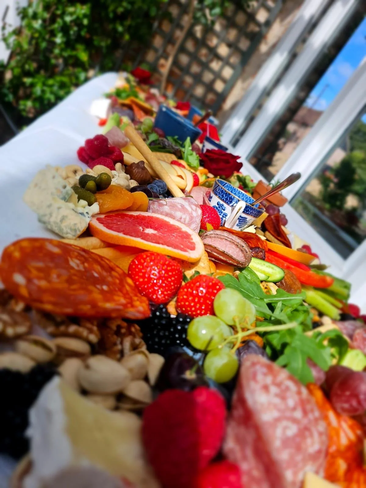 A long table set with a variety of cheeses, fruits, vegetables, meats, and snacks, outdoors on a patio with a white tablecloth and string lights.