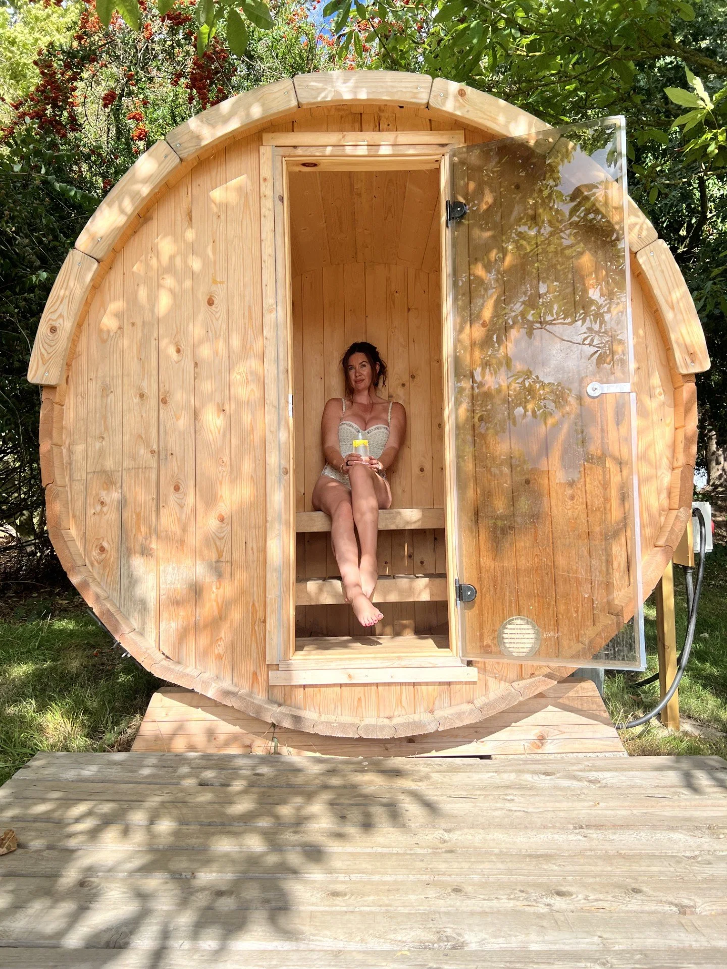 A woman sitting inside a small wooden barrel-shaped sauna with an open glass door, surrounded by green foliage and trees.