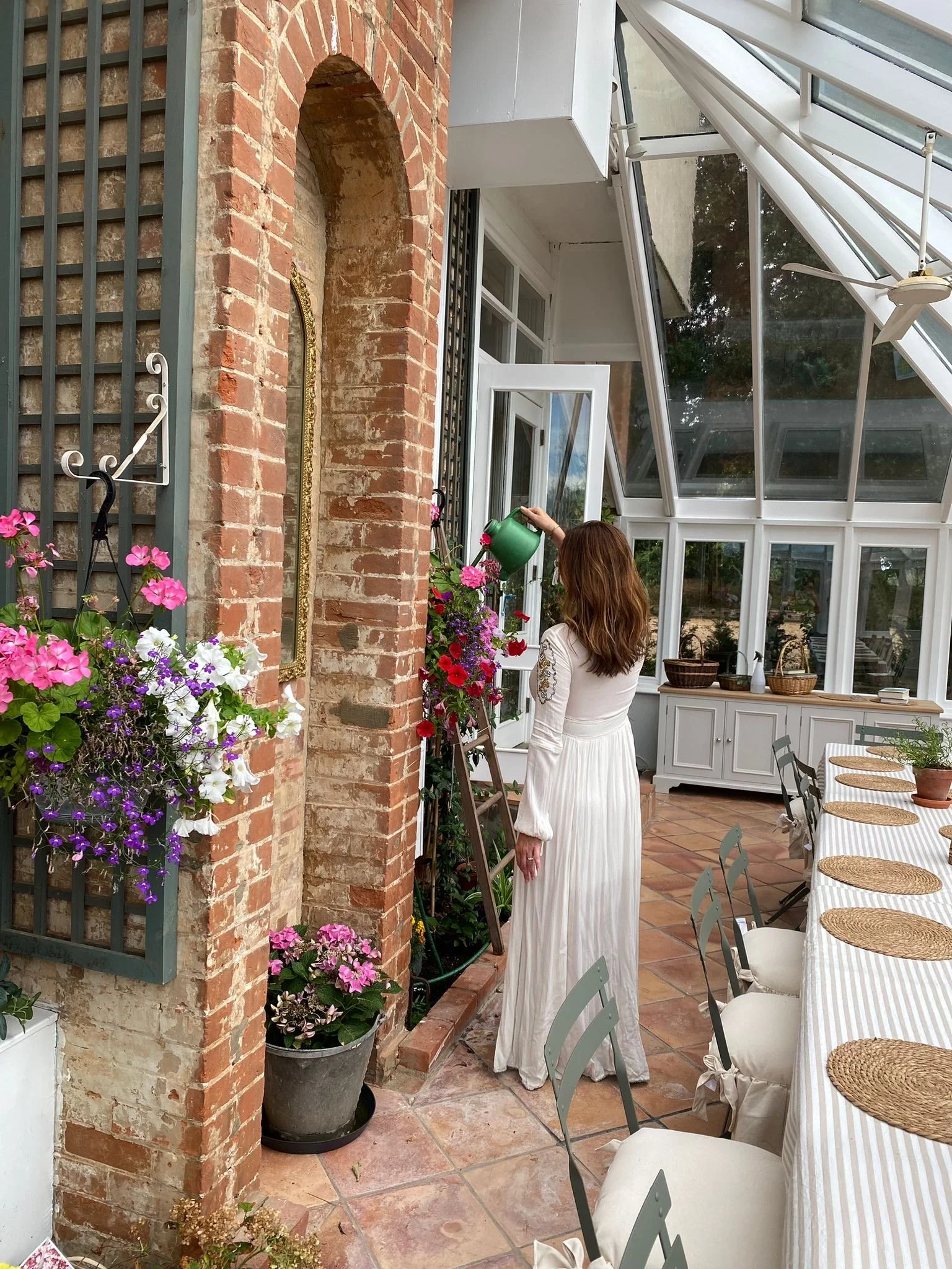 Woman watering hanging and potted flowers inside a greenhouse attached to a brick wall, with a long dining table set with woven placemats and chairs.