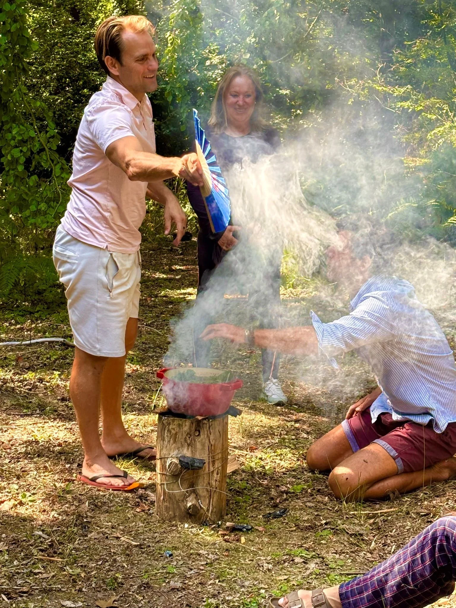 People gathered outdoors in a wooded area; one man is standing and pouring a blue bag into a pot over a fire, while a woman stands behind him smiling, and another person is kneeling and reaching into the pot amidst smoke. setting intentions