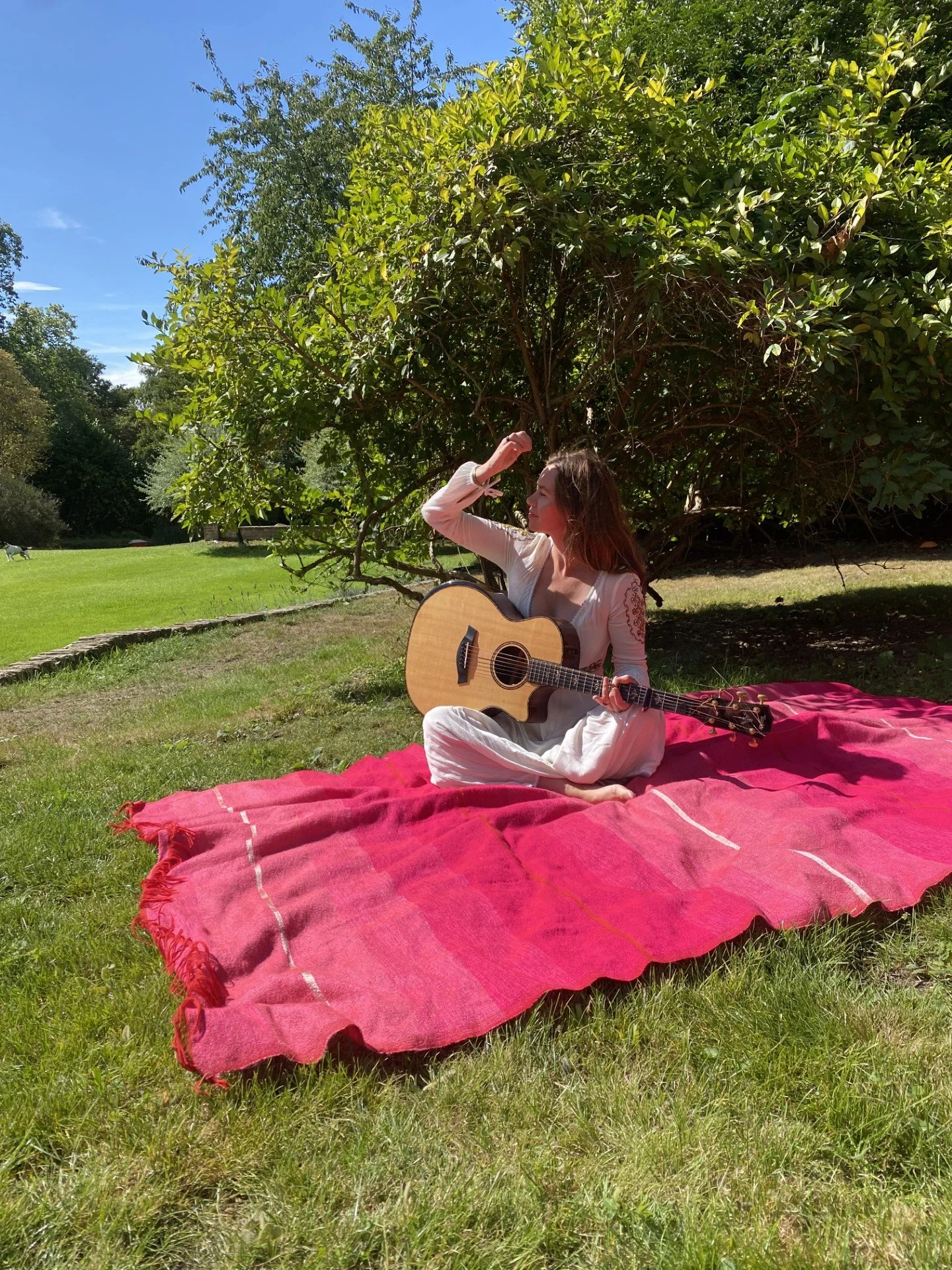 A woman sitting on a red blanket outdoors, holding an acoustic guitar, with a large green bush and trees in the background under a clear blue sky.