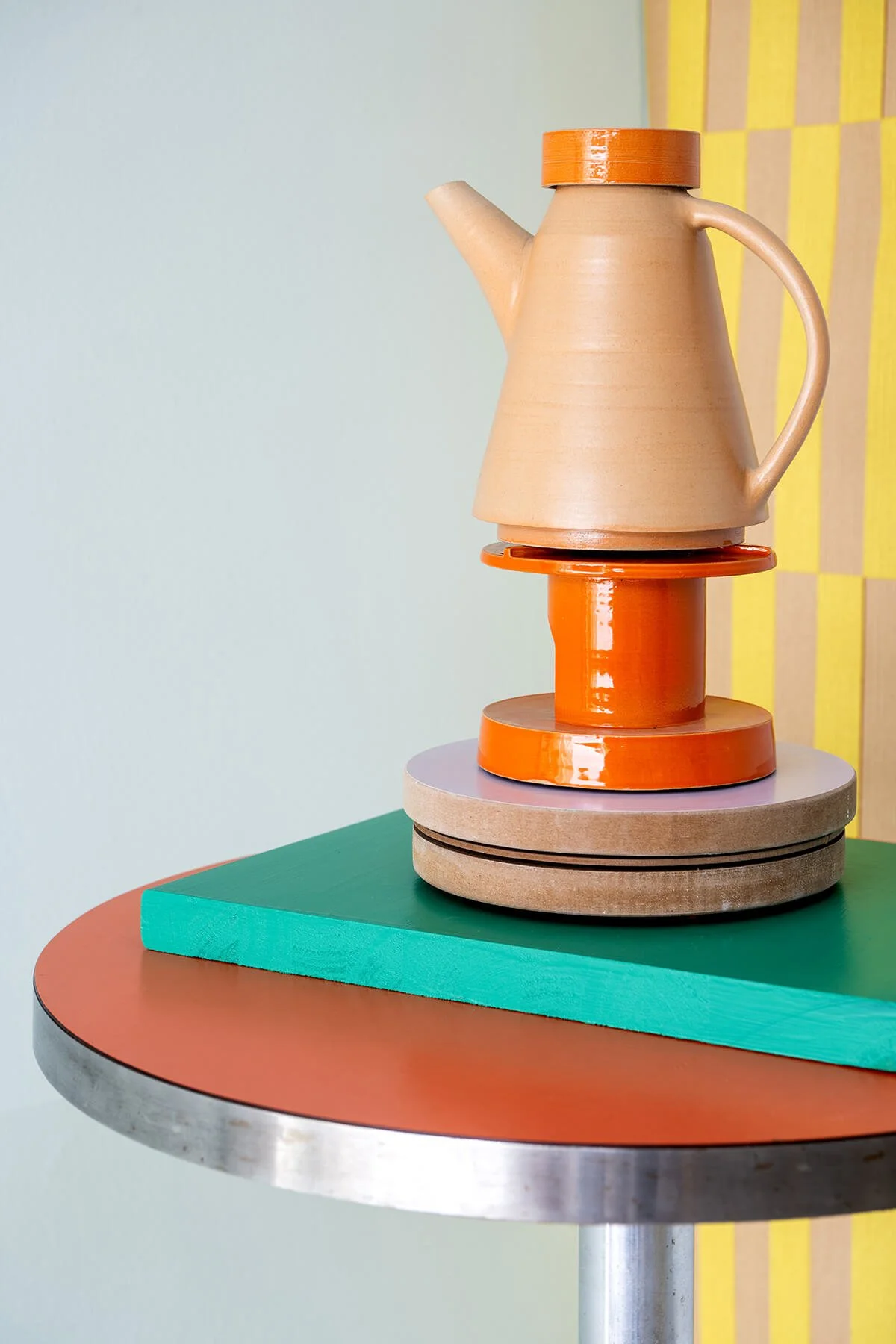 Handgemaakte steengoed theepot met oranje rechaud, gefotografeerd in een kleurrijke interieursetting op een oranje en groene tafel.