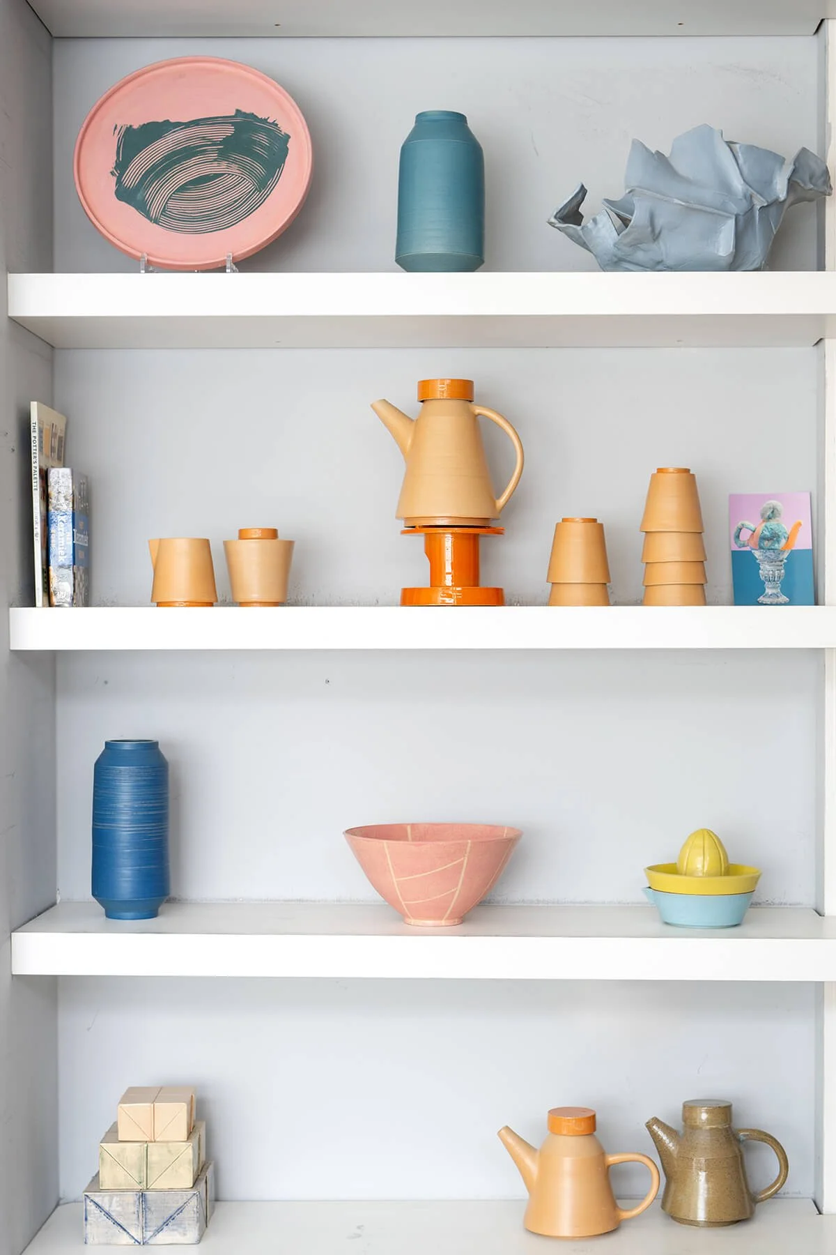 Handgemaakte keramiekcollectie van LAB L.L. op een witte wandkast, met vazen, kommen, citruspers en kannen in roze, blauw, geel en oranje tinten.