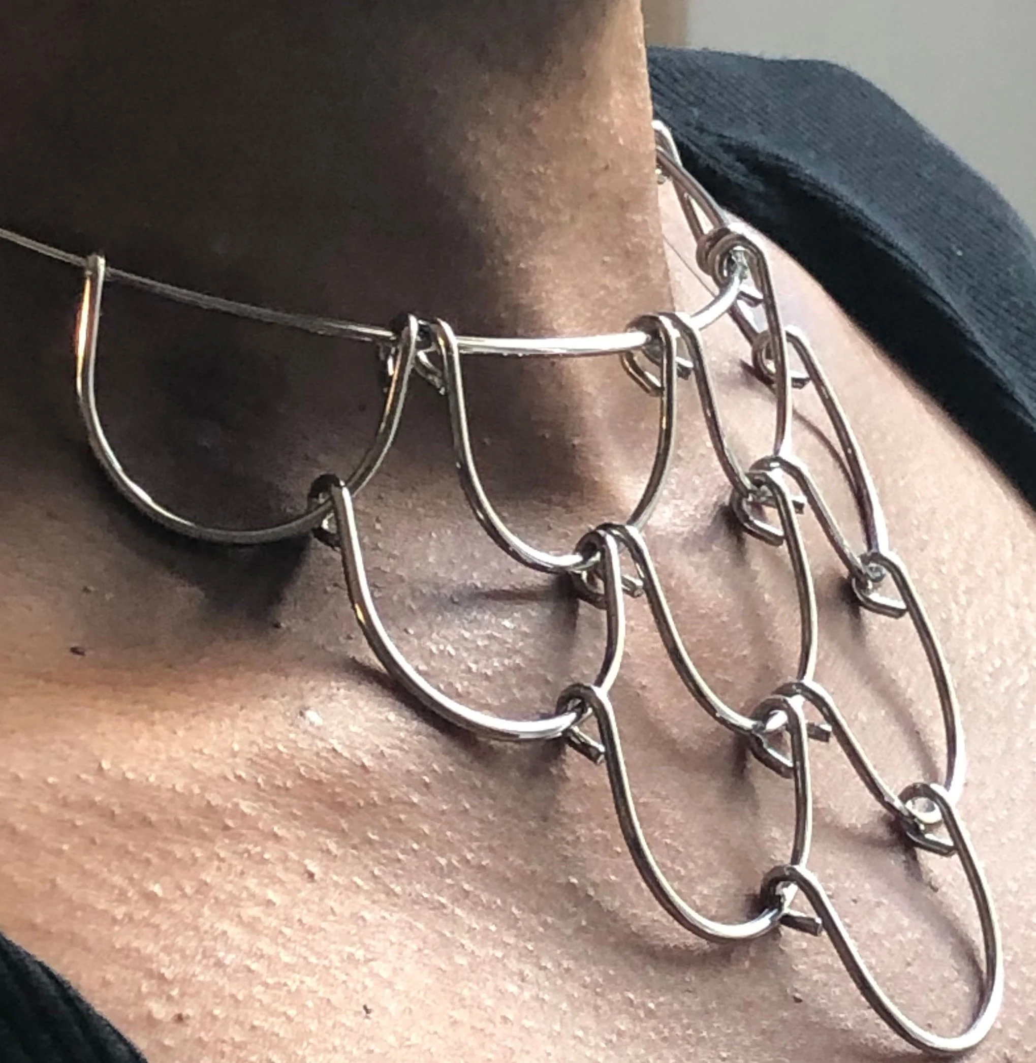 Close-up of a person's neck wearing a unique, layered silver chain necklace with large, interlinked loops and small hooks, paired with black sunglasses.