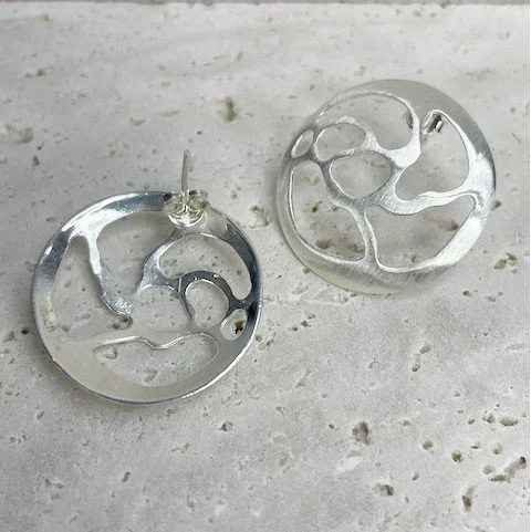 Silver circular earrings with abstract cut-out designs placed on a light-colored surface.