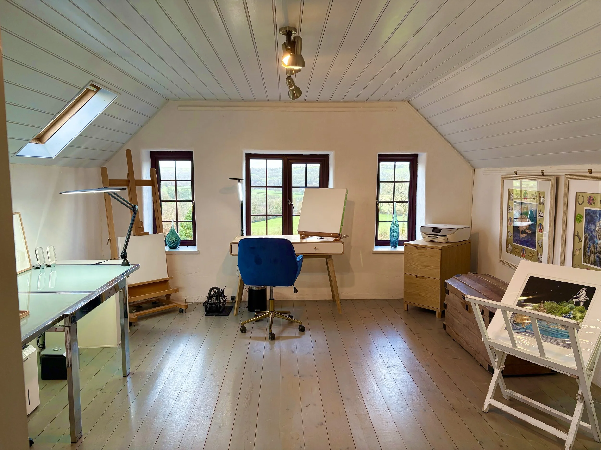 Attic art studio with three windows, a sketchboard, blue chair, wooden drawers, a printer, and artwork on the walls. Natural light from the windows and skylight.