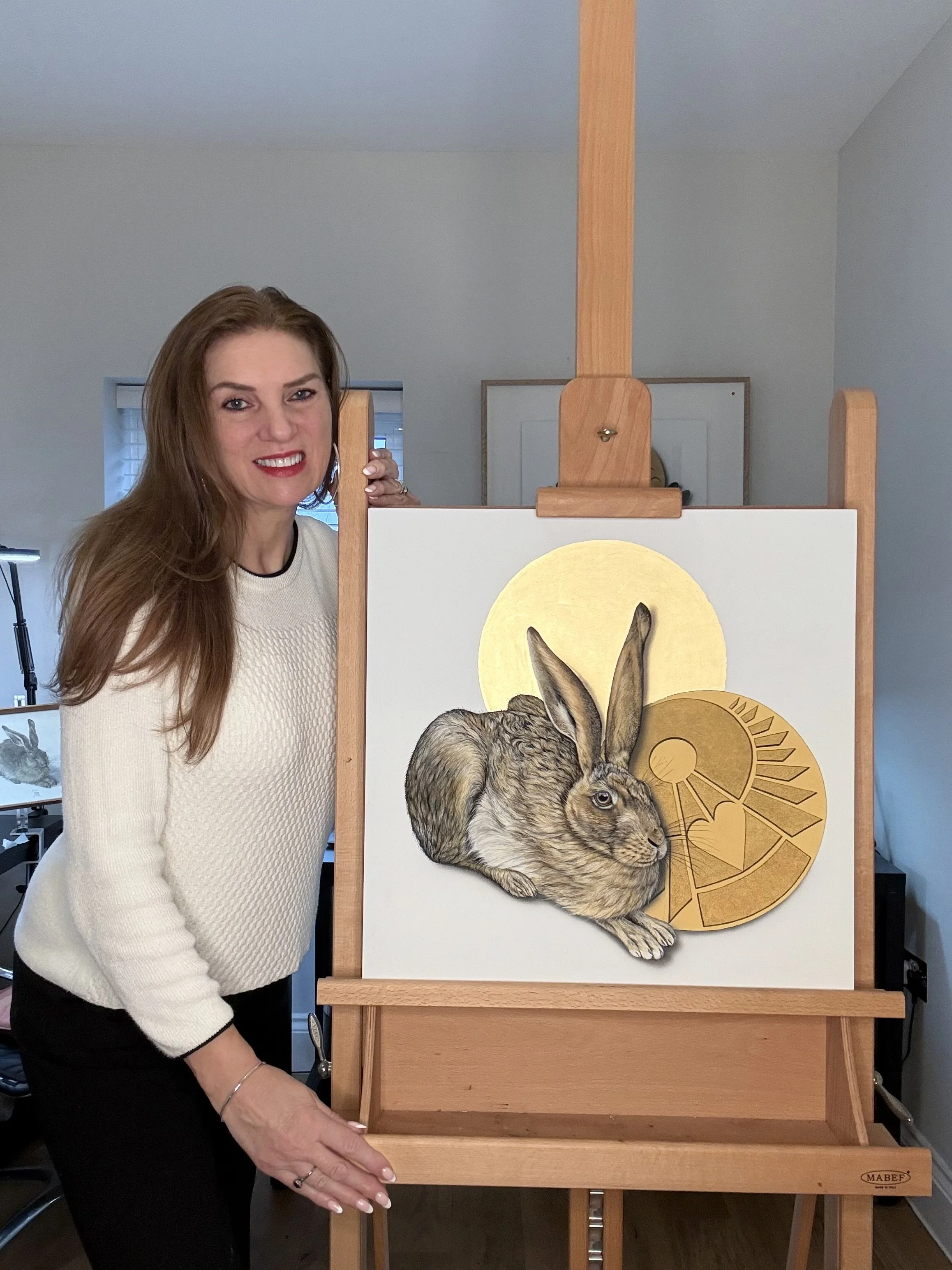A woman with long hair, wearing a white sweater, standing next to an easel with a painting of a rabbit with large ears, gold circles, and geometric patterns in gold and black.