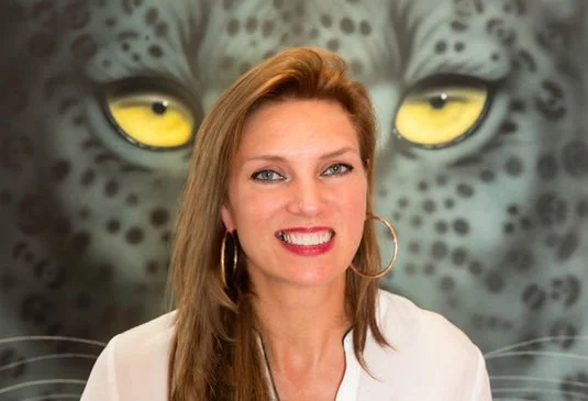 A woman with light brown hair and hoop earrings smiling in front of a large leopard face mural.