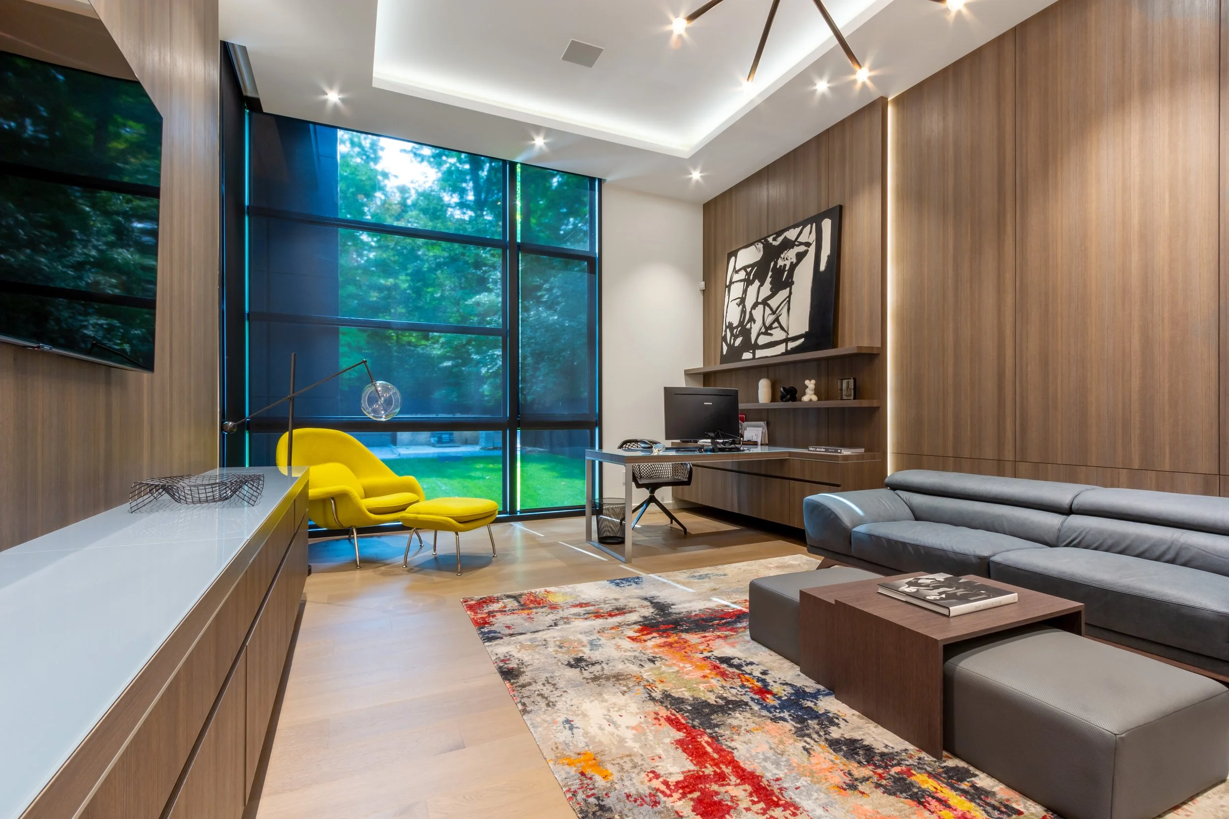 Modern office with wood-paneled walls, a large window with shades, a gray sofa, a colorful area rug, a yellow chair with a matching ottoman, a desk with a computer, and contemporary artwork.