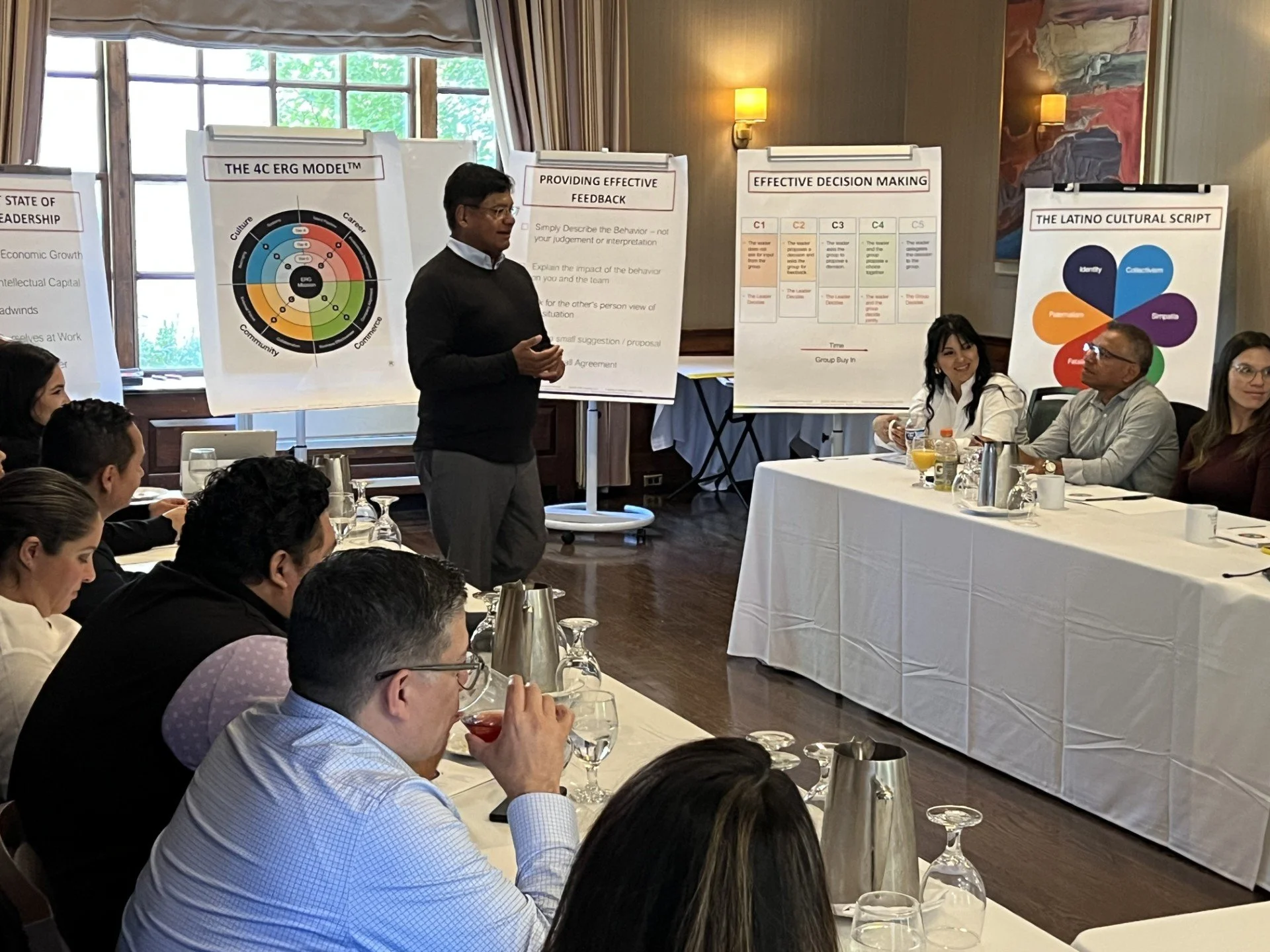 A group of professionally dressed people seated at tables in a conference room listening to a presenter. The presenter is standing near posters about feedback, decision making, and cultural scripts, with one poster titled "The 4C ERG Model." The room
