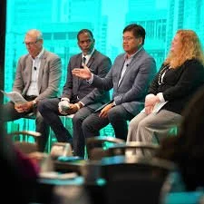 Four people sitting on a panel at a conference or event, with a blue cityscape background.