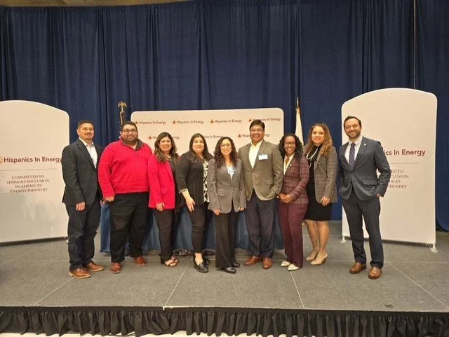 Group of nine diverse individuals standing on a stage at a conference, with a backdrop that reads 'Hispanics in Energy' and related text.