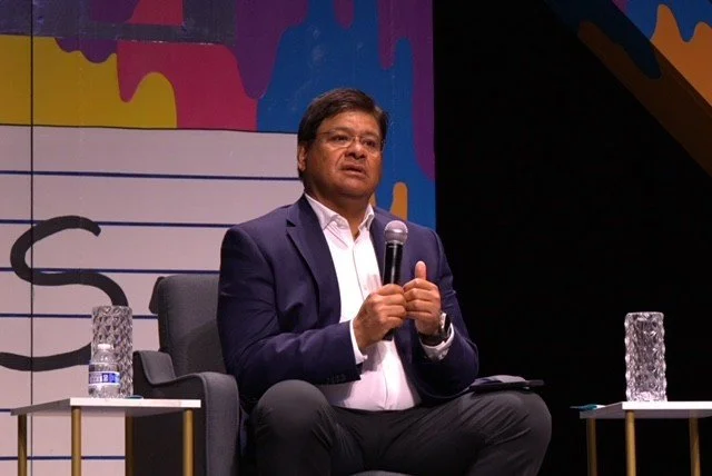 A man in a navy suit and white shirt sitting on a stage, holding a microphone, with two glasses of water on small tables beside him.