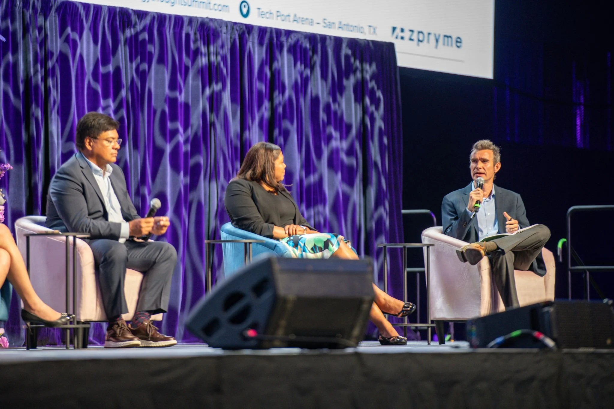 Four panelists seated on stage at a conference, with one man speaking into a microphone. The background features a purple curtain and a screen displaying conference details.