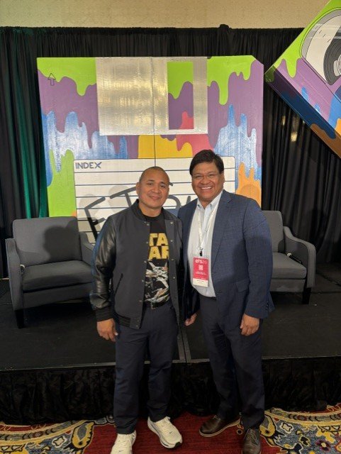 Two men smiling and posing for a photo on stage with colorful backdrop and black couches in the background.