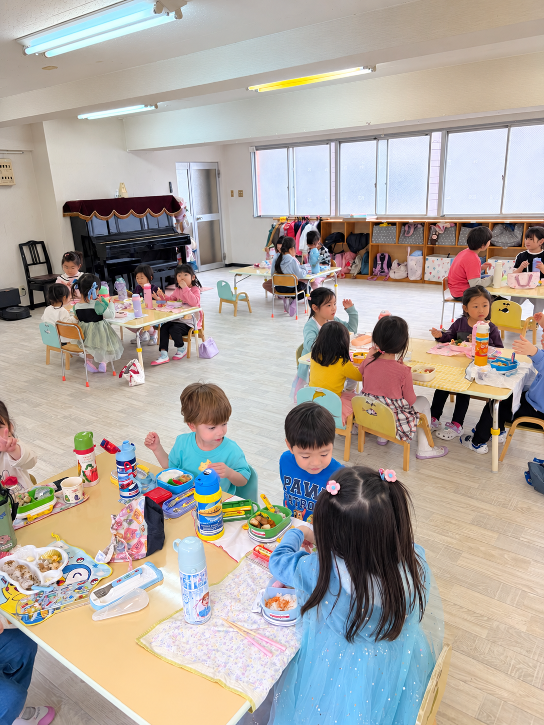 幼稚園の子供たちがお弁当を食べている教室の様子。子供たちはテーブルに座り、友達と話しながらお弁当を楽しんでいる。背景にはピアノと靴箱がある明るい教室です。