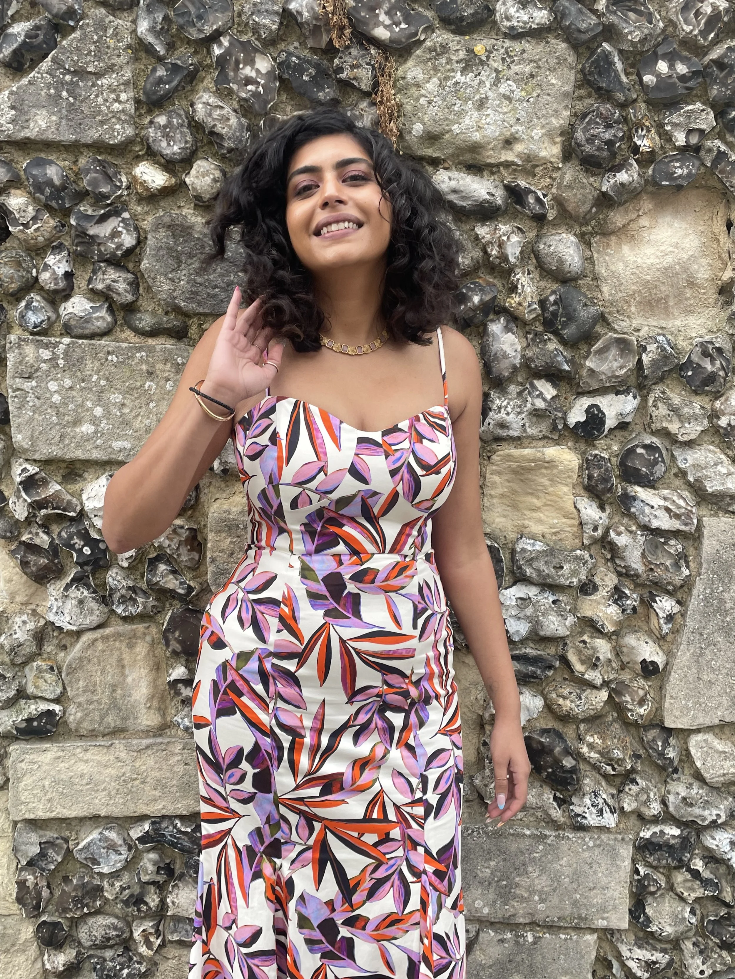 A woman with curly black hair wearing a colorful, patterned dress standing against a stone wall.