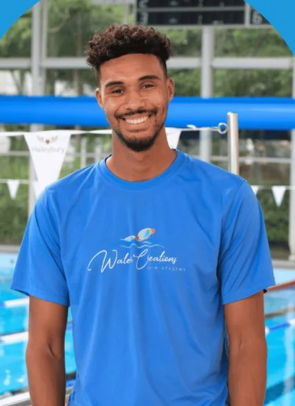 A young man with curly hair and a beard smiling, wearing a blue Water Academy T-shirt, standing near a swimming pool with a sports facility in the background.