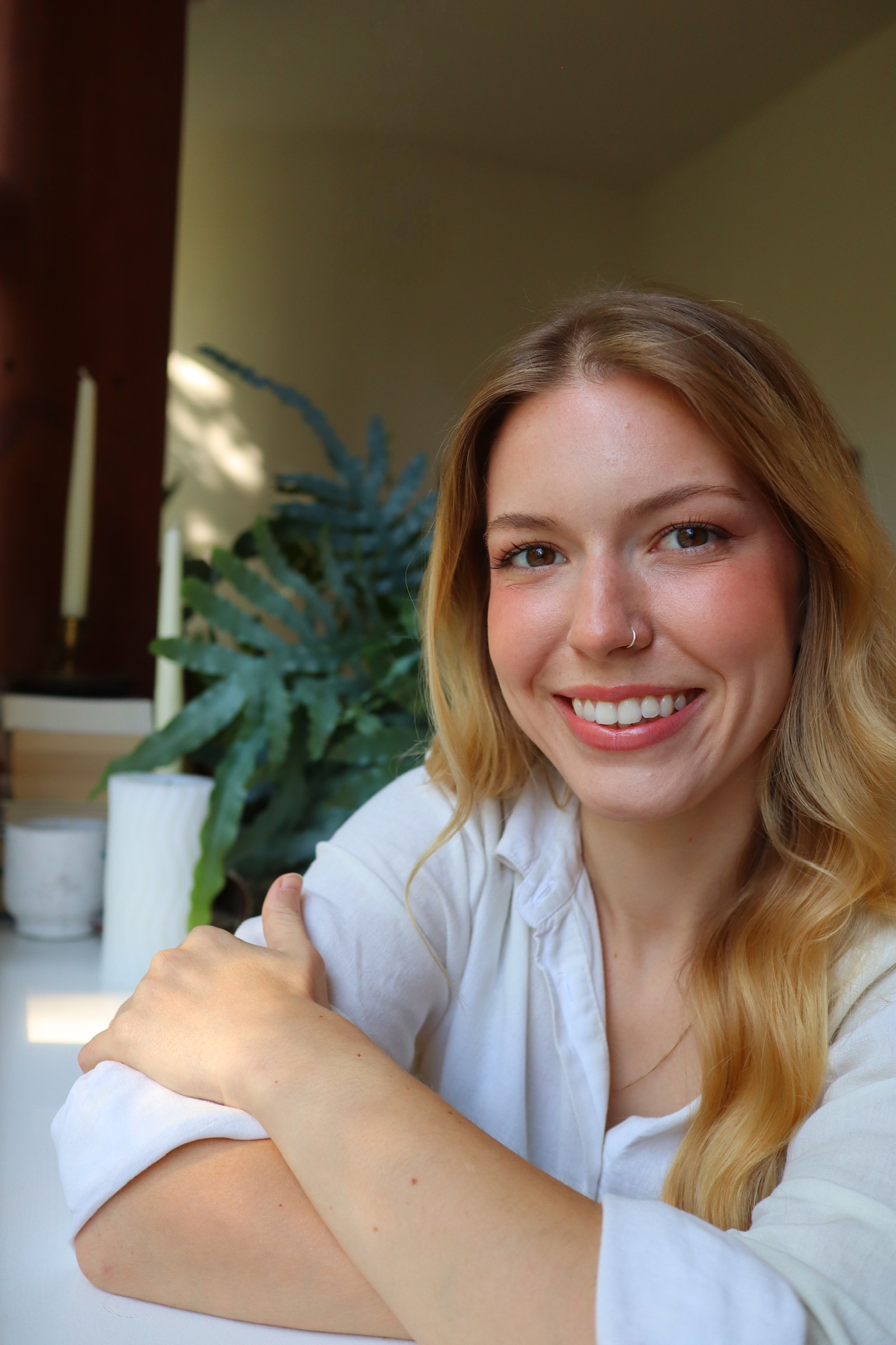 A smiling woman with blonde hair and a nose piercing, sitting indoors with her arms crossed, in front of a green plant and candles.