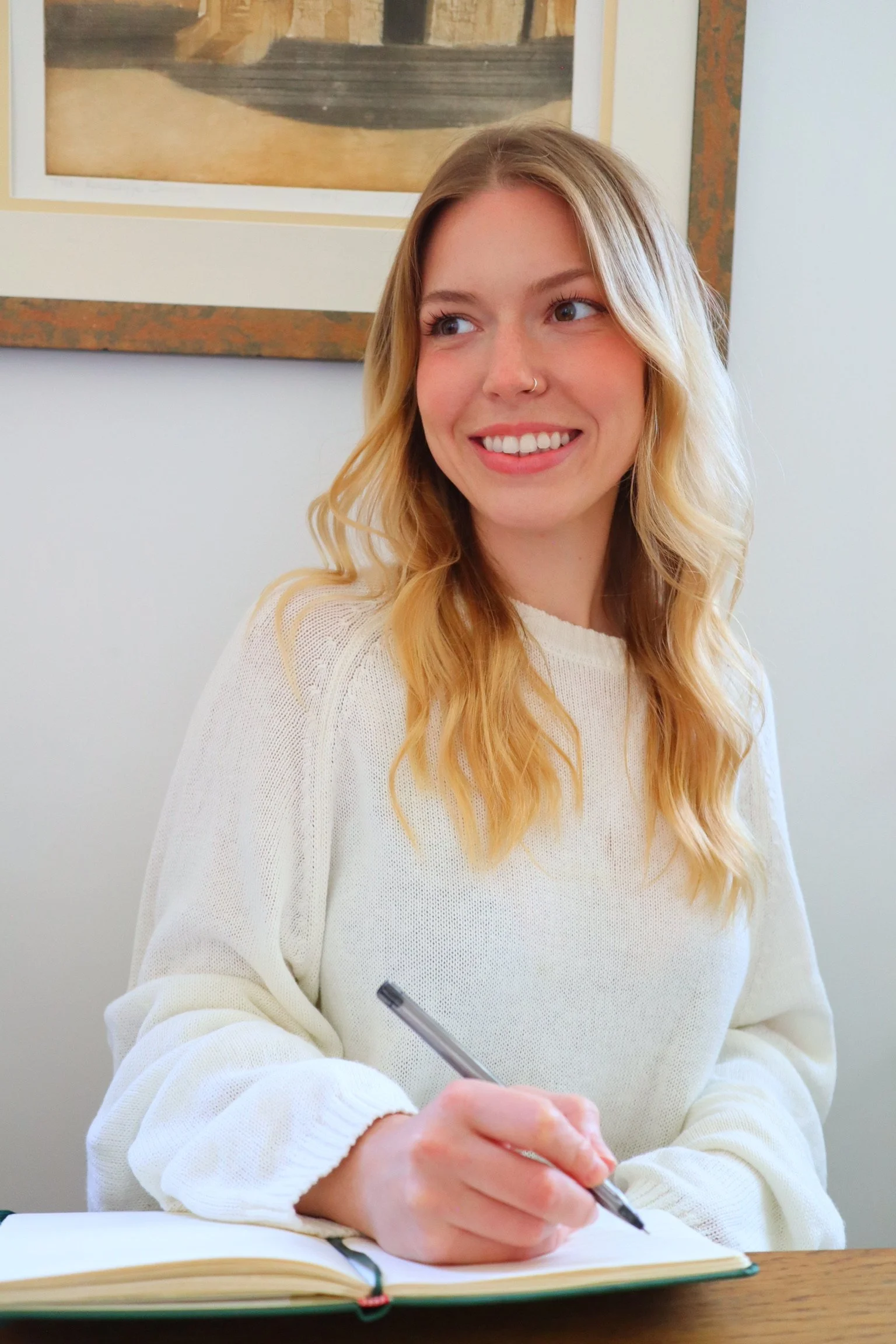 A young woman with long blonde hair, smiling, wearing a cream-colored sweater, sitting at a desk with an open notebook and holding a pen, in a room with a framed painting on the wall.