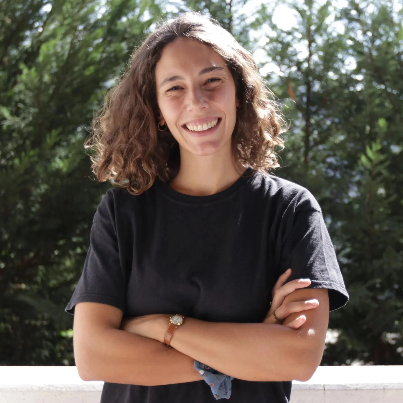 Una giovane donna con capelli ricci e sorriso, indossa una maglietta nera e braccialetti, si trova all'aperto con uno sfondo di alberi verdi.