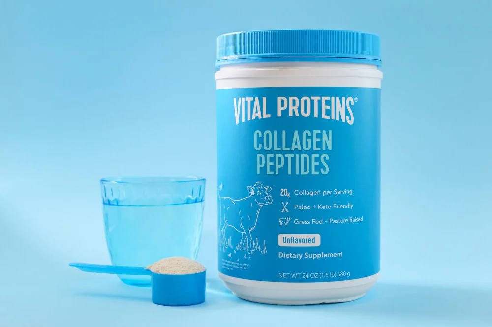 Container of Vital Proteins collagen peptides supplement, a glass of water, and a scoop of powder on a blue background.