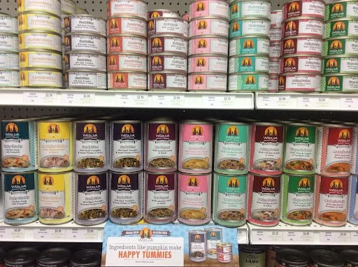 Shelves filled with cans of Wysong dog food in various flavors, including turkey, beef, and vegetable, with a box of Happy Tummies pumpkin and sweet potato dog treats at the bottom.