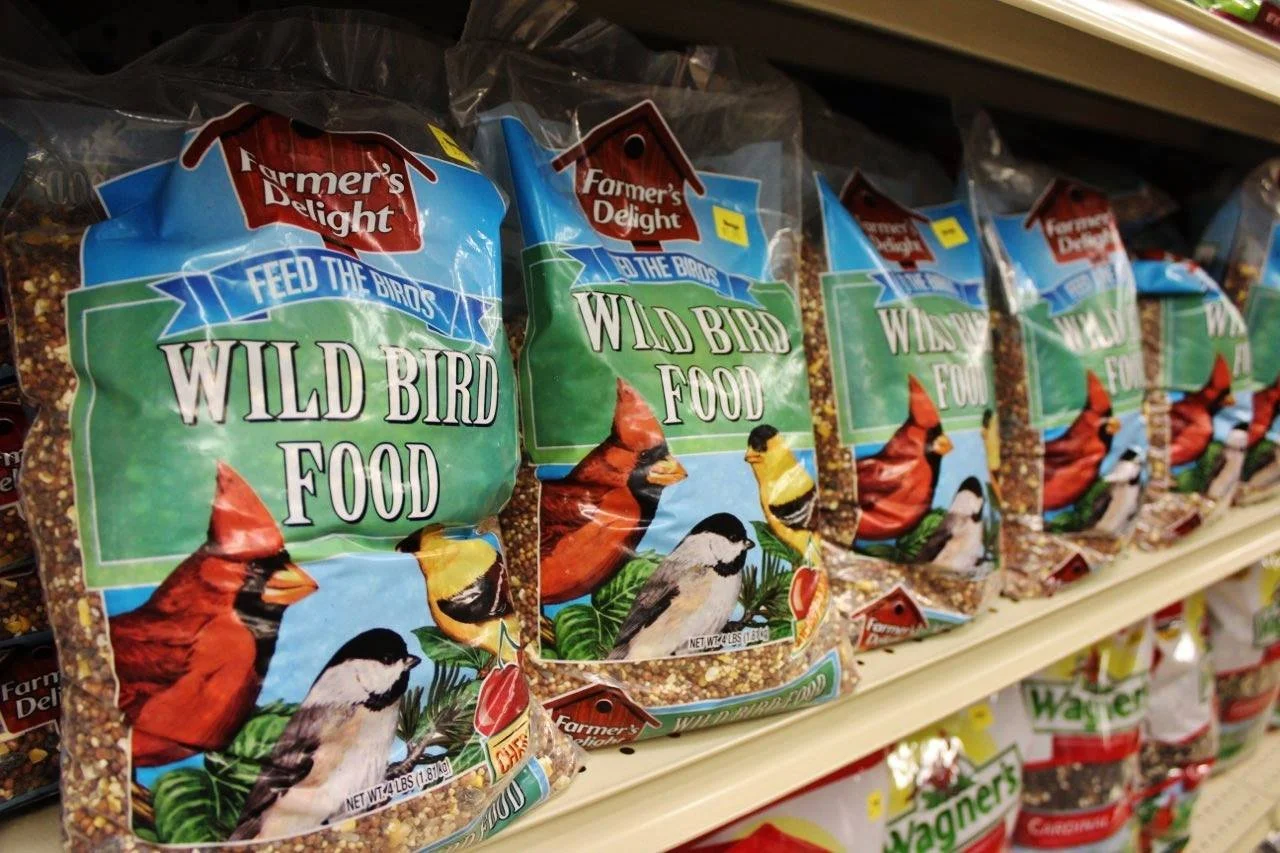 Packages of bird seed on store shelf labeled 'Wild Bird Food' from Farmer's Delight, featuring illustrations of cardinal, goldfinch, and chickadee birds, with the text 'Feed the Birds.'